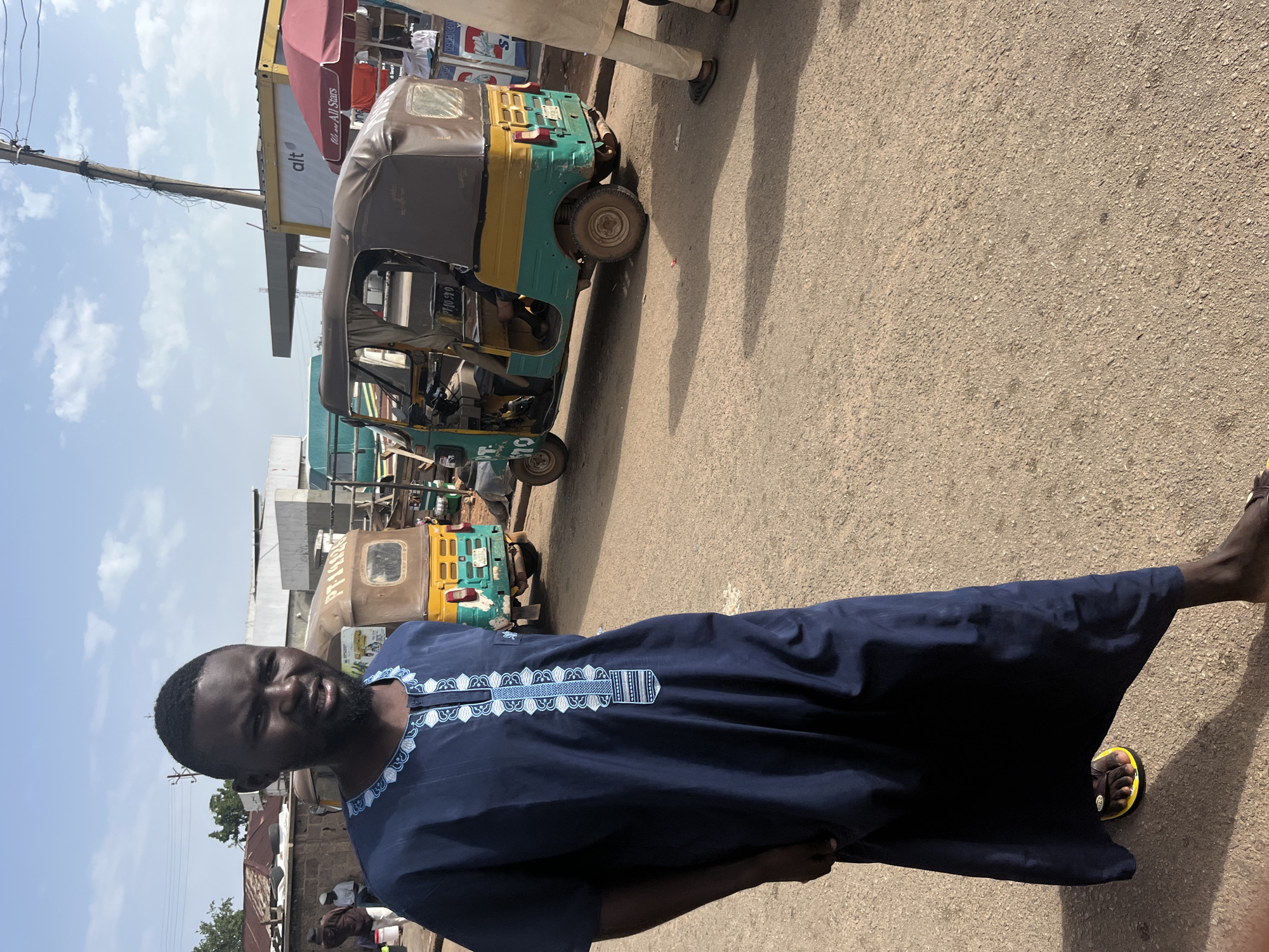 In Gura Top, Nigeria, a man stands on a dusty road in front of a row of three-wheeled vehicles. He wears a dark blue robe with intricate white and blue embroidery around the neckline and down the front. The man is looking towards the camera, with a slight smile and his mouth open as if speaking. Behind him, several auto-rickshaws, also known as Keke-NAPEPs, are parked or in motion. One is a vibrant green and yellow, with a driver visible. Another, predominantly brown and yellow, is positioned further back. The environment appears to be a marketplace or transportation hub, with some stalls and buildings visible in the background under a bright, clear sky. The road is unpaved and appears to be made of packed dirt and gravel.
