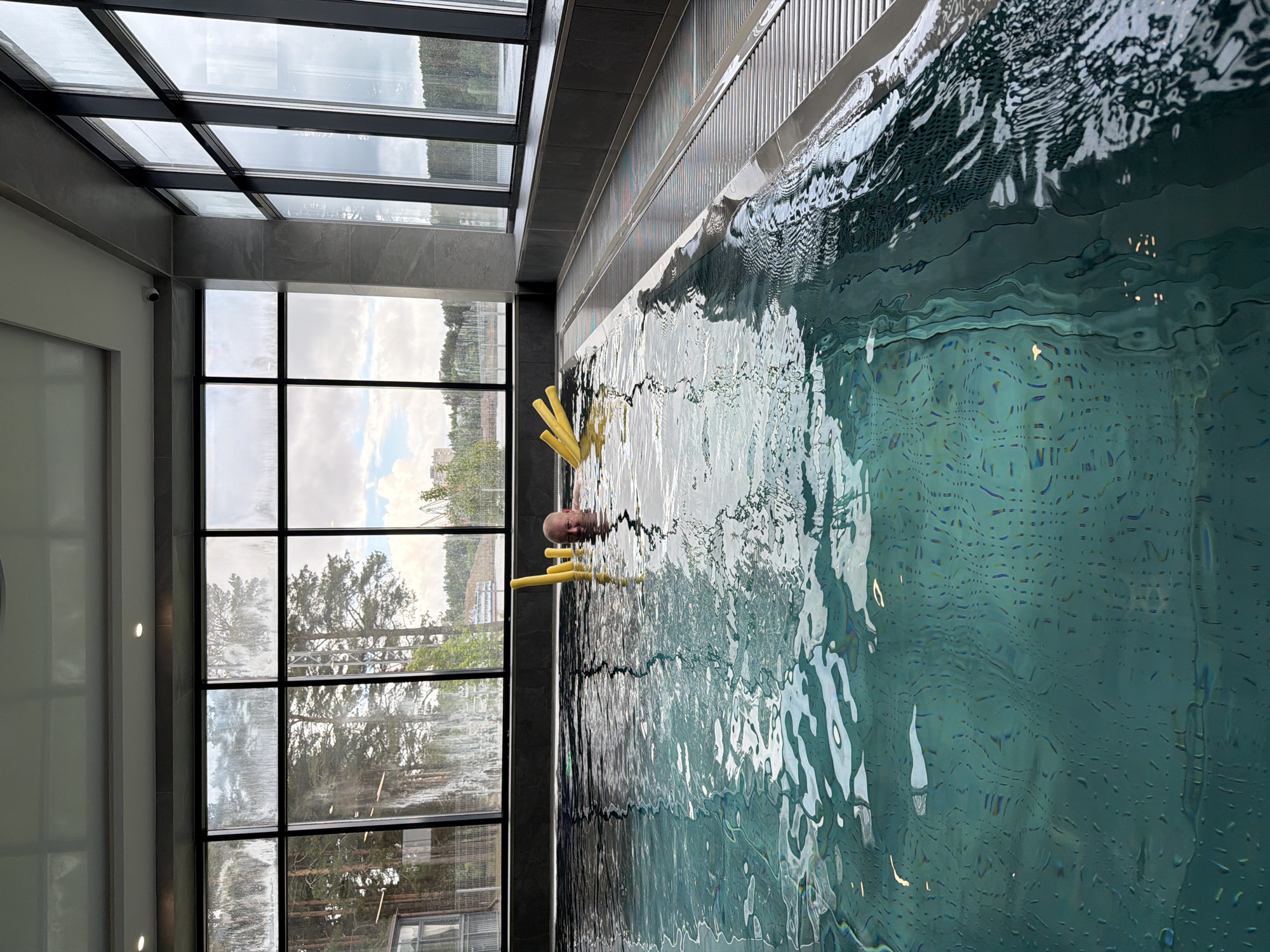 A man is in a swimming pool at the "Chaika" sports center in Chaikovsky, Russia. The man is holding yellow pool noodles and is near the edge of the pool. Large windows beside the pool show a view of trees and buildings. The pool water is light turquoise with many reflections. The building's structure includes a combination of windows, metal grids, and tiled surfaces.
