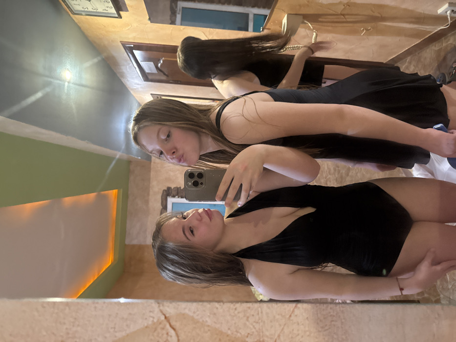 The image captures a mirror selfie featuring two young women indoors. The scene is brightly lit by internal lighting, and the presence of reflections suggests a large mirror.
In the foreground, a young woman with long, light brown hair stands on the right side of the frame, holding a modern smartphone (with a triple camera lens visible) in her left hand, positioned to capture her reflection and the reflection of another person. She is wearing a black, low-cut, possibly one-piece swimsuit or bodysuit. Her expression is neutral, and she is looking towards the phone's camera. Her right arm is bent, with her hand resting near her hip.
Behind and slightly to the left of the first woman, the reflection of a second young woman is visible. She also has long, light brown hair and is wearing a black top or dress with thin straps. She is making a playful "puckered lips" or "kissing" face towards the camera. Further back in the reflection, what appears to be another view of this second woman (or a third person) is visible, leaning over a surface, with long dark hair cascading down.
The background of the room, as seen in the mirror's reflection, displays varied interior design elements. The ceiling is a dark blue-grey, featuring a bright, recessed light fixture that creates a starburst effect. A section of the wall on the left side of the reflection is a light green or teal color, illuminated by a strip of warm, orange-yellow light, possibly LED backlighting. The wall on the right side appears to be a light brown or beige, with a textured finish, possibly mimicking stone or stucco.
Several objects are visible in the background reflections: an analog clock with hands is mounted high on a wall, though the exact time is not legible. Below it, a vintage white rotary phone with a coiled cord rests on a surface. A wooden doorframe is also discernible in the background, along with a window showing a glimpse of blue sky or distant buildings. The overall ambiance is warm and casual, typical of a personal setting.