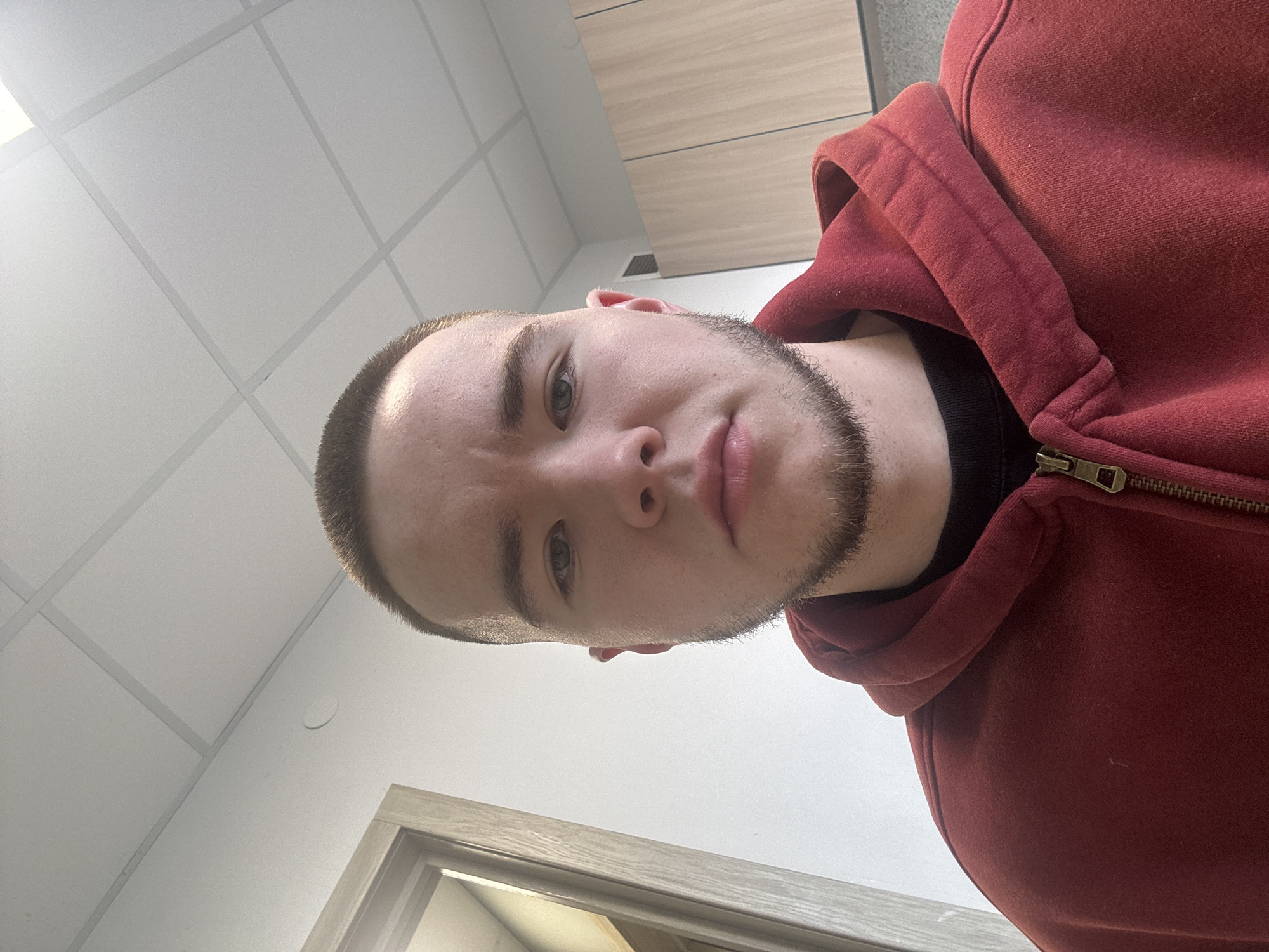 A man with short brown hair and a beard is wearing a red hooded sweatshirt. He is looking at the camera. The background is an indoor setting with a white ceiling and walls. A door is visible in the lower left corner. The location is not discernible from the image.