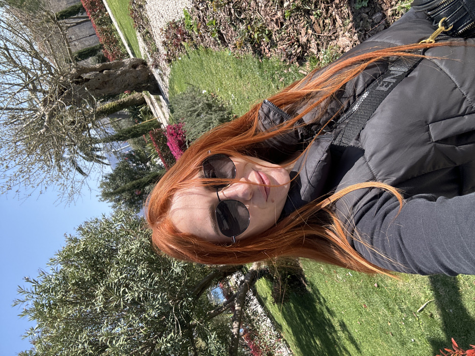 A female subject with long, auburn hair, wearing sunglasses, a black long-sleeved shirt, and a black puffer vest, is depicted taking a selfie outdoors under a clear blue sky. A black shoulder bag with a visible "ENI" inscription on its strap is across her body. The background features a well-maintained park or garden with green grass, cultivated flower beds displaying pink/magenta blooms, and various trees including a large leafy green tree, a bare-branched tree, and a palm tree. Distant water is visible through foliage on the left. Stone architectural structures, characteristic of the Old Town, are discernible behind the trees, indicating the location is within a park adjacent to the Old Town of Budva, Montenegro.