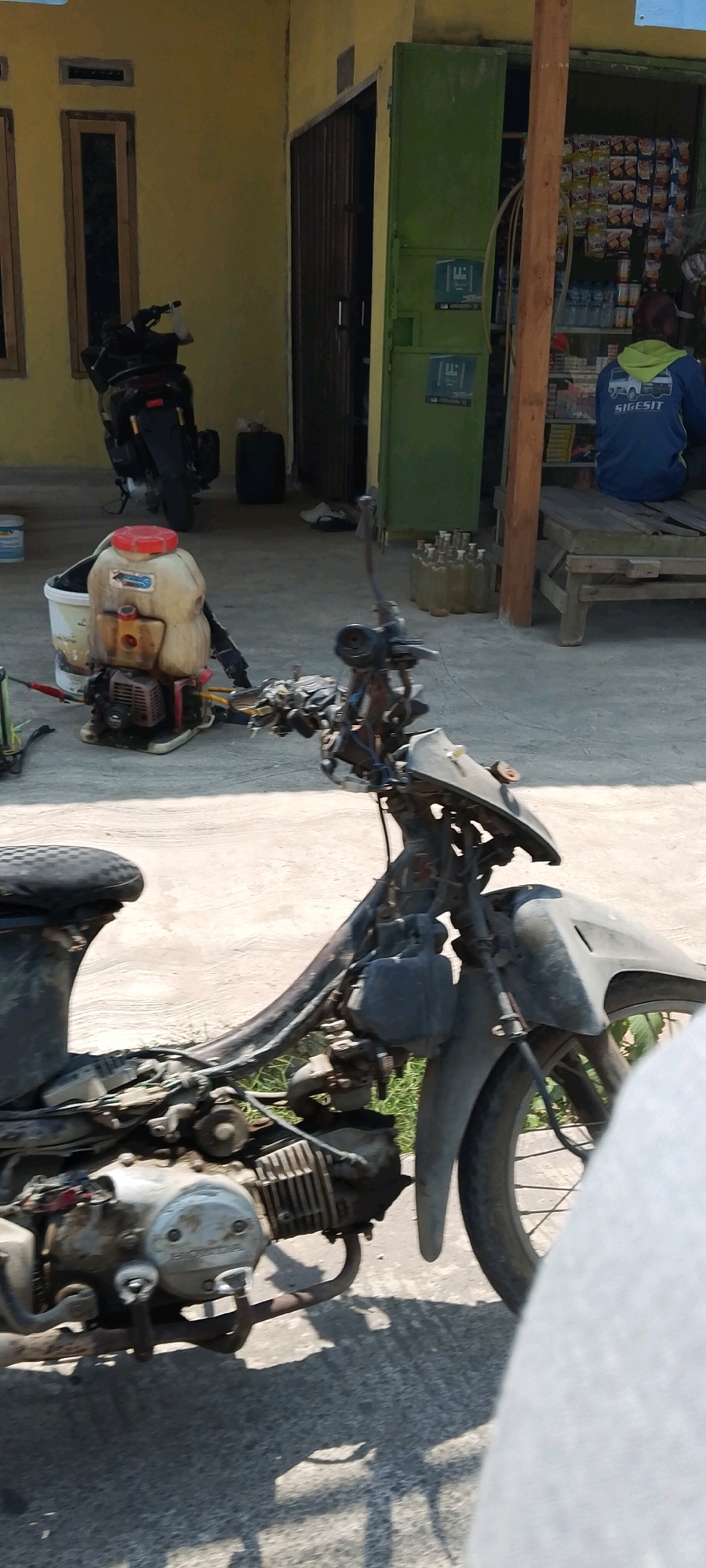 In a city like Banjarsari, Indonesia, a scene unfolds that is both common and indicative of daily life. The primary subject is a worn-out motorcycle, its frame exposed, hinting at disrepair or ongoing maintenance. This motorcycle is parked on a concrete surface, possibly a sidewalk or yard, in front of a building with a yellow facade. To the left of the motorcycle, a weathered pesticide sprayer with a red cap sits on a white bucket. Behind the sprayer, a dark-colored motorcycle is partially visible, leaning on its stand.
The building itself features a green metal door, and to its right, a small stall or shop is open, displaying various packaged goods on shelves. A person, seen from the back, is seated on a wooden bench in front of the shop. They are wearing a blue hooded jacket with green accents and a graphic with the text "SIGESIT" on the back. Several glass bottles, likely containing liquid, are arranged in a row on the ground near the stall. The lighting suggests it is daytime, with shadows cast by the objects onto the ground. The overall environment appears to be a residential or commercial area, typical of a bustling Indonesian city.