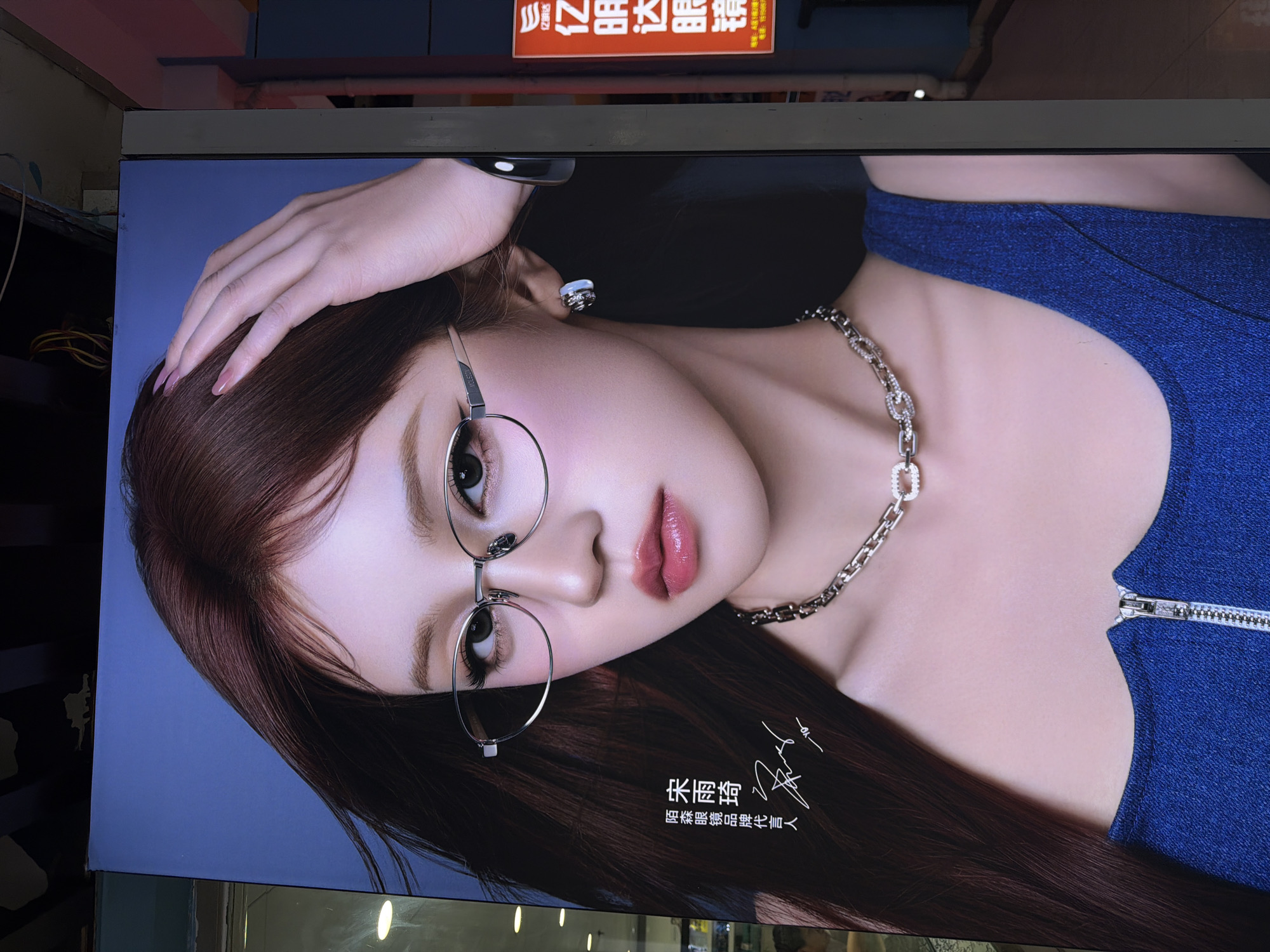 The image shows a large advertising billboard in Jiangning District, China. The billboard features a close-up portrait of a young Asian woman with brown hair, wearing glasses and a blue top with a silver zipper. She is also wearing a silver necklace and earrings. Her left hand is placed on her head. Below her image, in Chinese characters, is a name and a description indicating she is a spokesperson for a product. The billboard is lit from below. Overhead, other signs and architectural details of the urban environment can be seen.