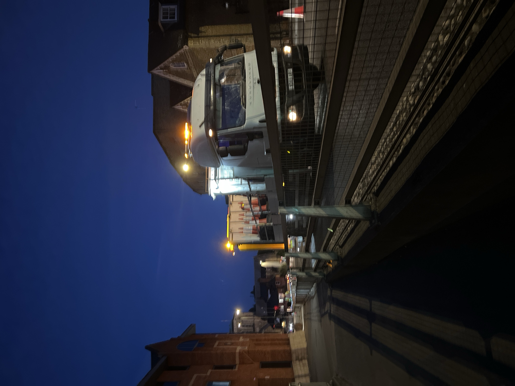 In the city of Guildford, United Kingdom, on an evening with a deep blue twilight sky, a white truck with an illuminated amber beacon is parked on a street. The truck, labeled "EUROCARGO", has its headlights on and appears to be carrying traffic cones in its bed. In the foreground, a dark paved walkway is visible, bordered by a wooden and metal fence. Shadows from the fence stretch across the walkway, creating a striped pattern. To the left of the walkway, a red brick building with arched windows is present. Further down the street, other buildings and streetlights are discernible, suggesting an urban environment. The scene implies ongoing road work or maintenance activities.