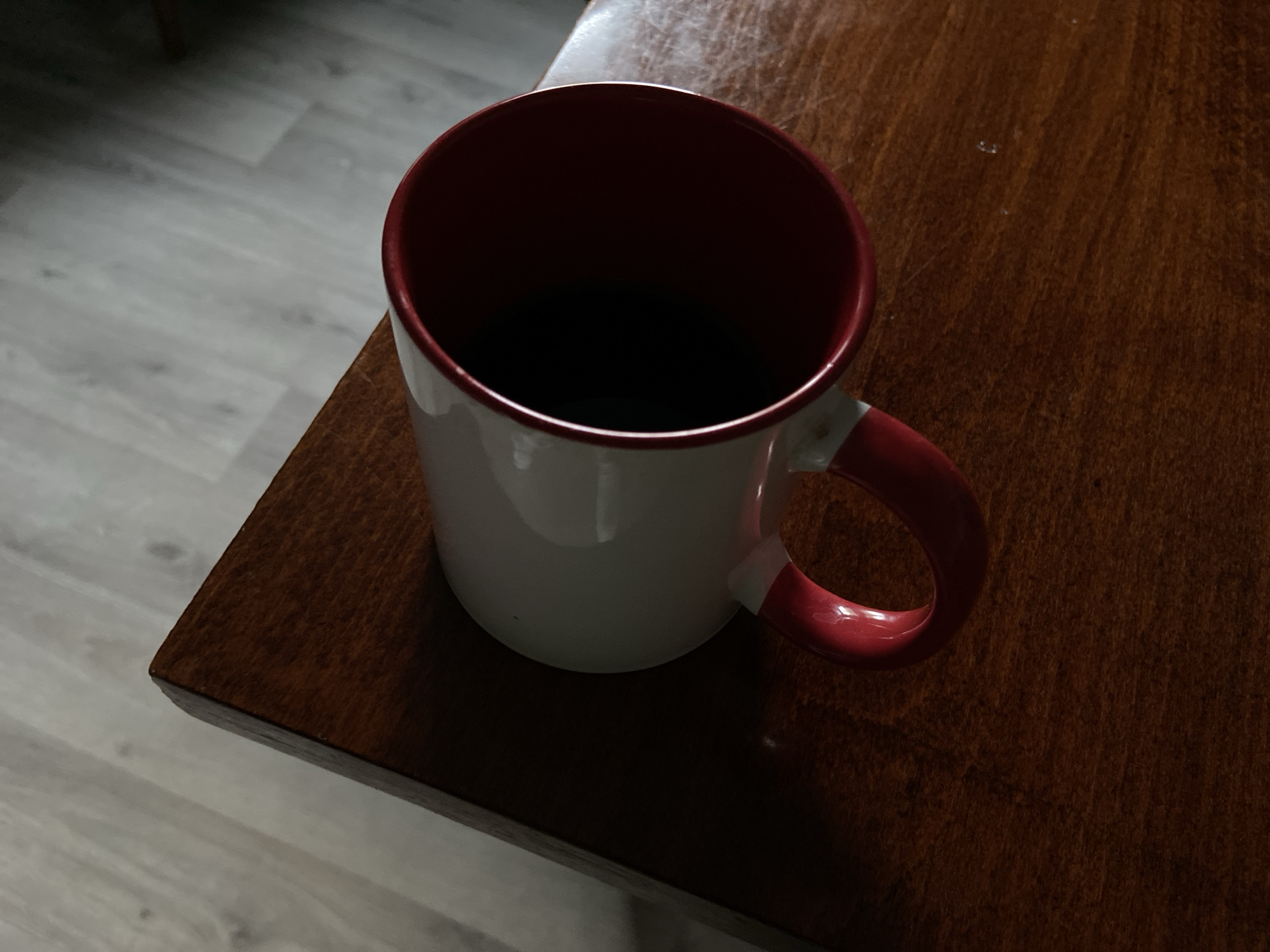 A white mug with a red interior and handle, filled with a dark liquid, sits on the corner of a polished wooden table. The table has a visible grain pattern and reflects some light. To the left of the table is a floor with light-colored wood planks. The lighting suggests an indoor environment, possibly a home or office, with shadows cast by the mug. There are no people or animals present in the image.