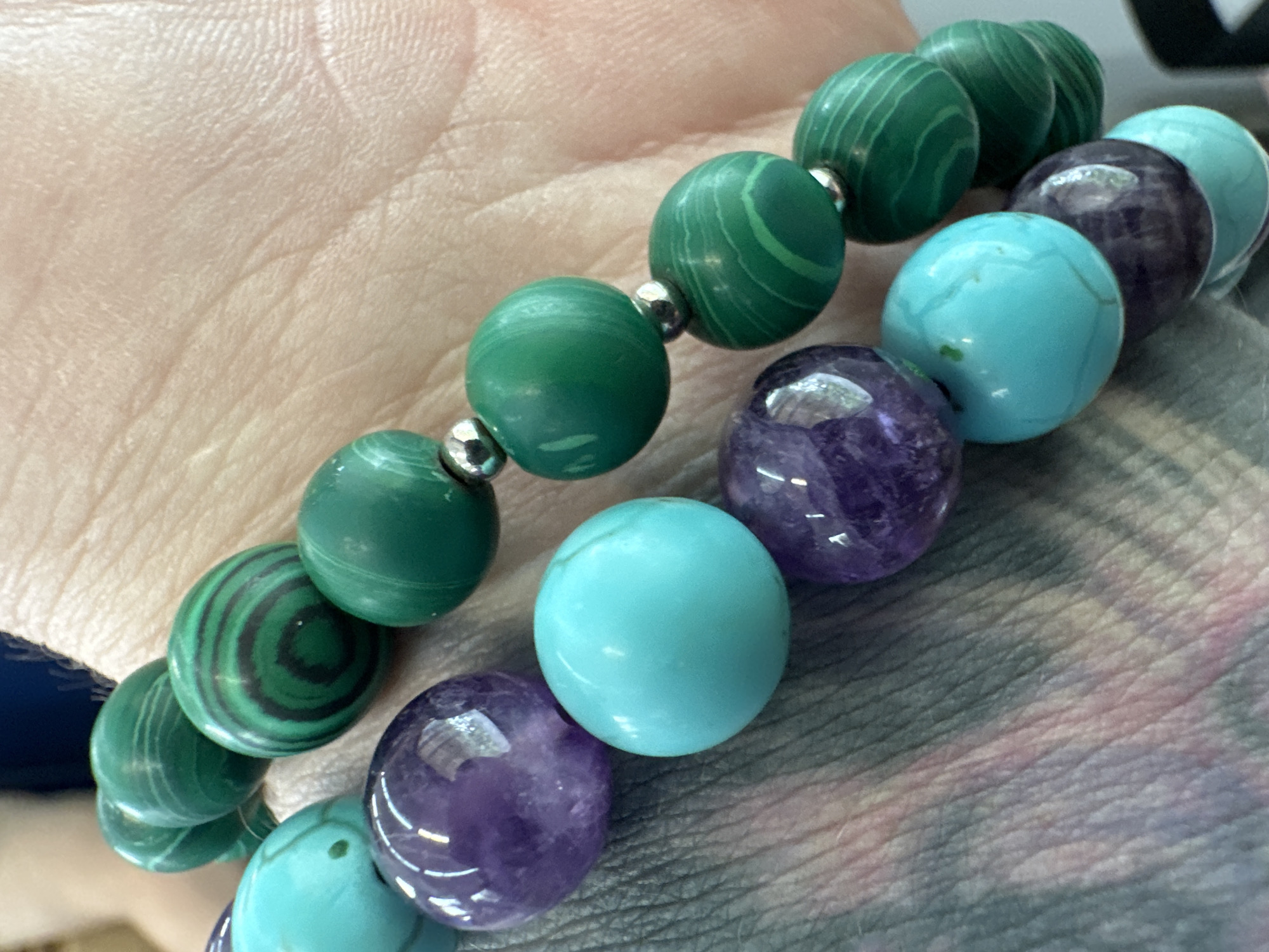 This is a close-up, overhead shot of two beaded bracelets worn on a person's wrist. The bracelets are composed of spherical beads of varying colors and patterns. One bracelet features vibrant green malachite beads with characteristic concentric banding, interspersed with small silver beads. The other bracelet alternates between translucent purple amethyst beads and bright turquoise beads. The skin of the wrist is visible, showing texture and light creases. The background is a blurry, textured surface that appears to be fabric or clothing. The context of Алабино, Russia is not discernible from the image itself.