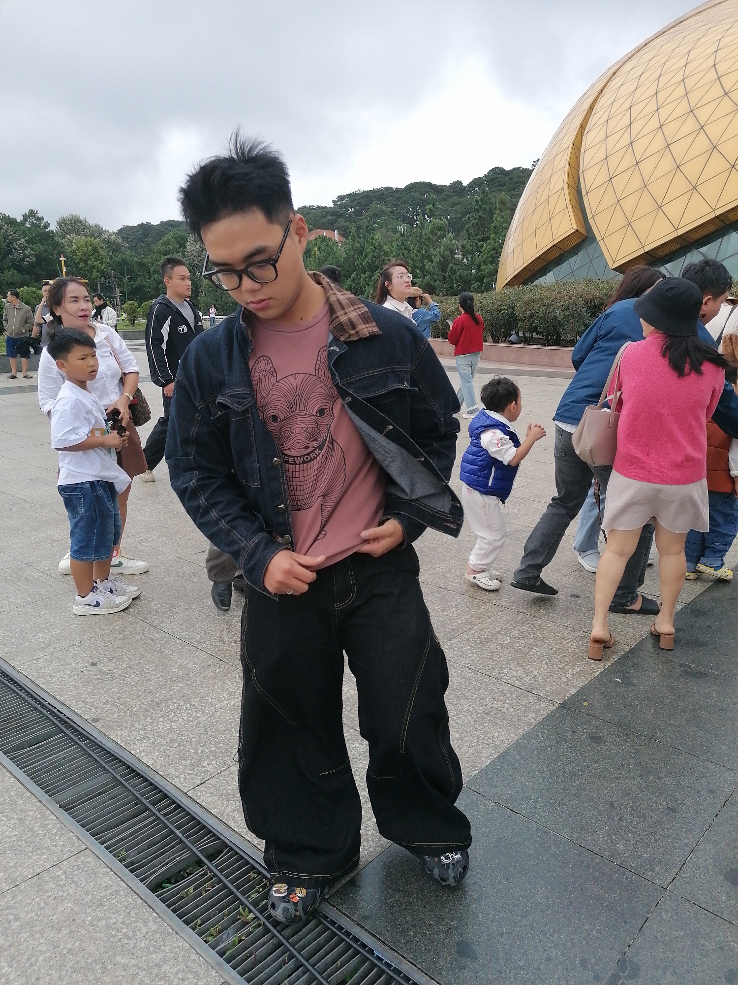 In Da Lat, Vietnam, a young Asian man with dark, spiky hair and glasses stands in the foreground, looking down. He wears a dark denim jacket over a pink t-shirt with a graphic of a dog's face and the word "LIFEWORK" on it. His pants are baggy and black. To his left, a young Asian boy in a white t-shirt and blue shorts stands with his mother, who is wearing a white top and holding a brown handbag. Behind them, other individuals are visible, including a man in a black jacket and a woman in a red jacket. In the background, a large, golden, dome-shaped building with a geometric pattern is prominent, surrounded by lush green trees. The ground is paved with stone slabs, and a metal grate runs along the left side of the frame. The sky is overcast.