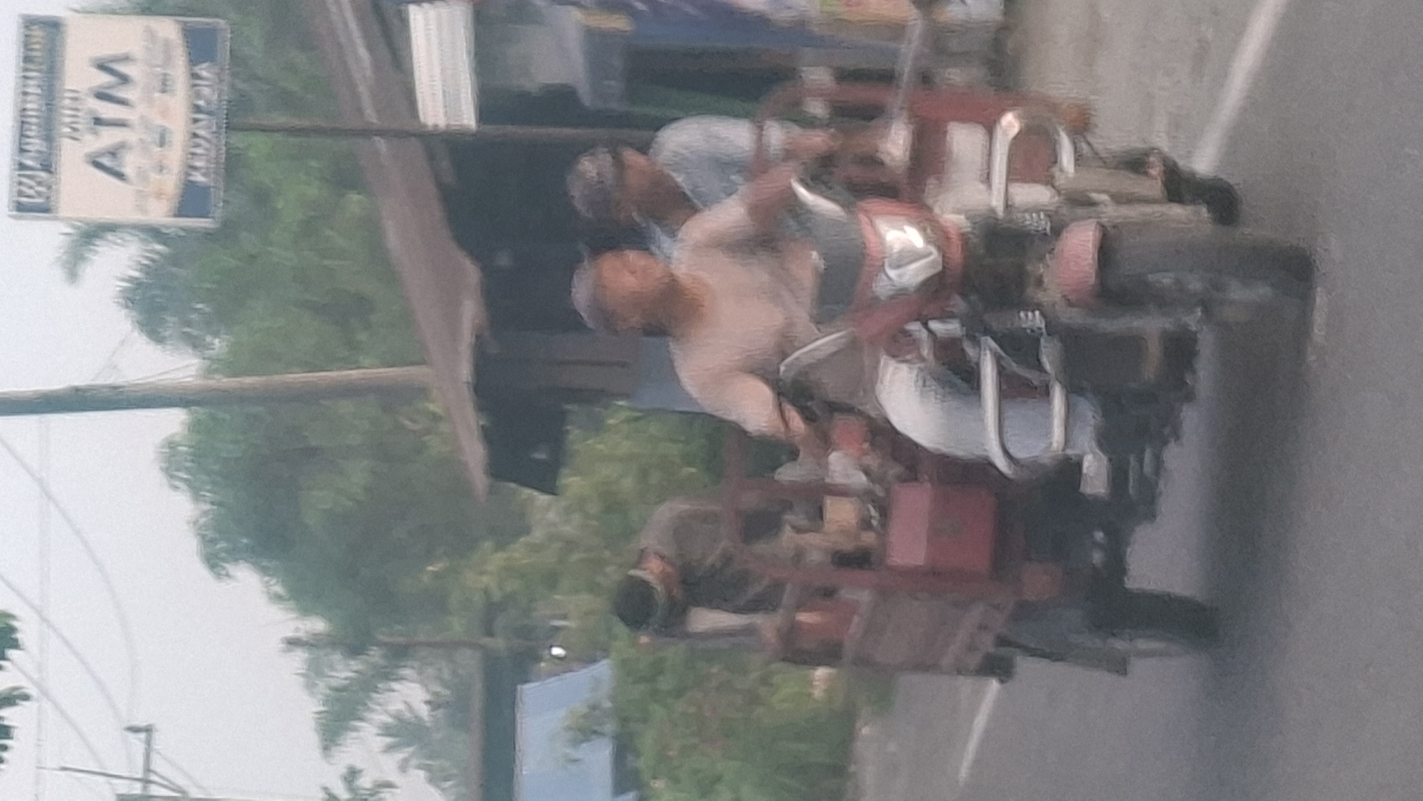 The image captures a street scene in Air Hitam, Indonesia. The central focus is a red motorized tricycle, commonly known as a "bajaj" or "bentor," carrying two individuals. The driver, wearing a light-colored t-shirt and a baseball cap, is positioned at the front, operating the vehicle. Behind him, another person is seated, also wearing a light-colored t-shirt and a cap. In the rear cargo area of the tricycle, a third person, dressed in darker clothing and wearing a head covering, is partially visible.
The tricycle is on an asphalt road. In the background, there are several structures, including a building with a weathered, corrugated metal roof. A utility pole with wires runs vertically through the scene. A sign for a "MINI ATM" is visible on a pole on the right side of the image. Lush green foliage and trees are present behind the structures, suggesting a semi-rural or suburban setting. The overall lighting suggests it might be daytime with overcast conditions. The image has a slightly soft focus, characteristic of a photograph taken with a mobile device in motion or low light.