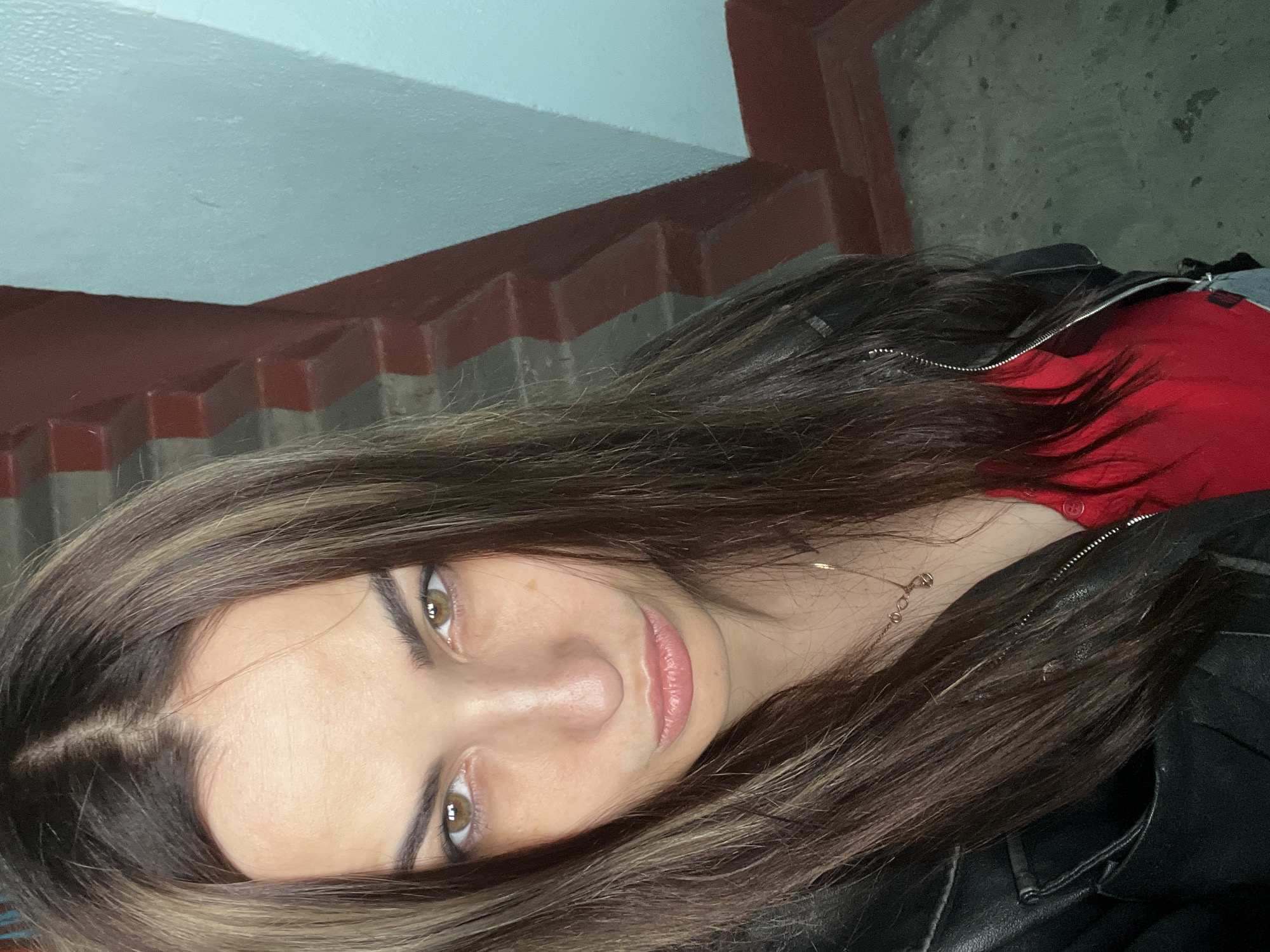 A female individual is depicted in a close-up, high-angle self-portrait within what appears to be an indoor stairwell or hallway in the city of Unknown, Unknown. She possesses long, dark brown hair with lighter brown streaks, parted centrally and framing her face. Her eyes are brown, and her eyebrows are dark. Her lips are slightly pursed in a pout. She wears a black leather jacket, partially unzipped, revealing a red garment underneath. A slender, gold-toned chain necklace is visible around her neck. In the background, to the subject's right, a structure with horizontally aligned reddish-brown elements, possibly stair treads or a decorative architectural feature, is present. Adjacent to this, a light blue wall section is visible. The floor in the lower right quadrant of the image consists of a dark, textured surface, possibly concrete, exhibiting minor discoloration.