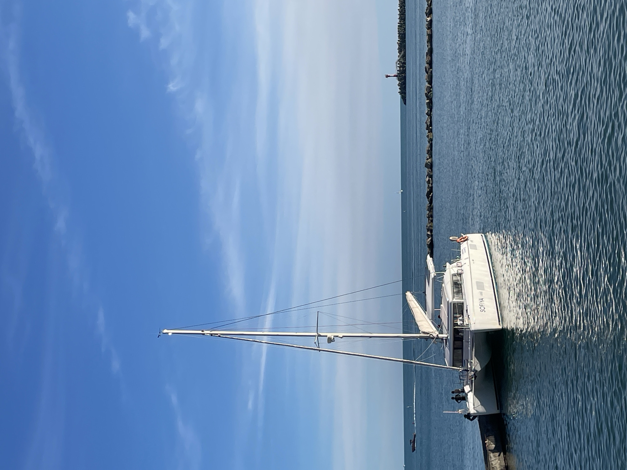 A white catamaran sailboat named "SOFIYA" is docked on the left side of the image. Three individuals, two appearing to be men, are visible on the deck of the boat. The catamaran is positioned against a dark, rough-textured pier. In the background, the calm, blue water stretches to the horizon under a bright blue sky with scattered white clouds. To the far right, a red and white lighthouse stands on a breakwater constructed of large, dark rocks. A few other distant boats are visible on the water, and a small sailboat is seen in the far distance to the left of the lighthouse. The scene depicts a serene coastal environment, likely within the city of Sochi, Russia, given the presence of a lighthouse and breakwater commonly found in port areas.