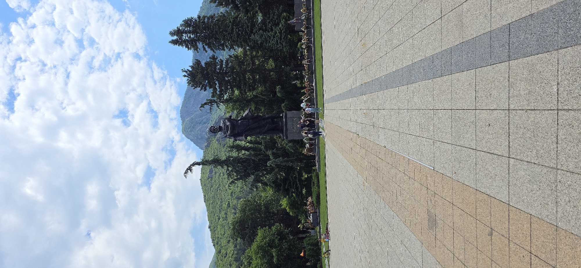The image displays a wide outdoor view of a monument situated in a public square, flanked by dense greenery and backed by mountains under a partly cloudy sky.
In the mid-ground, a large, dark-toned statue of a male figure stands on a stone pedestal. The figure has a mustache, wears what appears to be historical or military attire, holds a sword in his left hand, and has his right arm bent across his chest. Inscriptions are visible on the statue's base. Along the base of the statue, a continuous line of ceremonial floral wreaths is arranged on a strip of green grass. Several individuals are gathered near the statue; a group of approximately four people stands to the left of the base, and another group of three stands slightly to the right. A single person is seated on a low stone structure to the far right. On the far left, near the edge of the paved area and some trees, two children's bicycles, one red and one yellow, lie on the ground.
Lush, verdant trees and shrubs form a dense backdrop around and behind the monument. A prominent evergreen tree with a distinctively curved top stands to the left of the statue. Beyond the trees, a range of steep, green mountains rises, featuring visible rock formations and extensive tree cover. The sky above is bright blue with scattered white cumulus clouds.
The foreground is dominated by a large, paved plaza composed of square or rectangular light-colored tiles. A broad diagonal strip of light beige/tan tiles occupies the left side of the foreground, separated by a narrower dark grey diagonal strip from the main light grey tiled surface that extends across the rest of the plaza. The lighting indicates a bright, sunny day.
