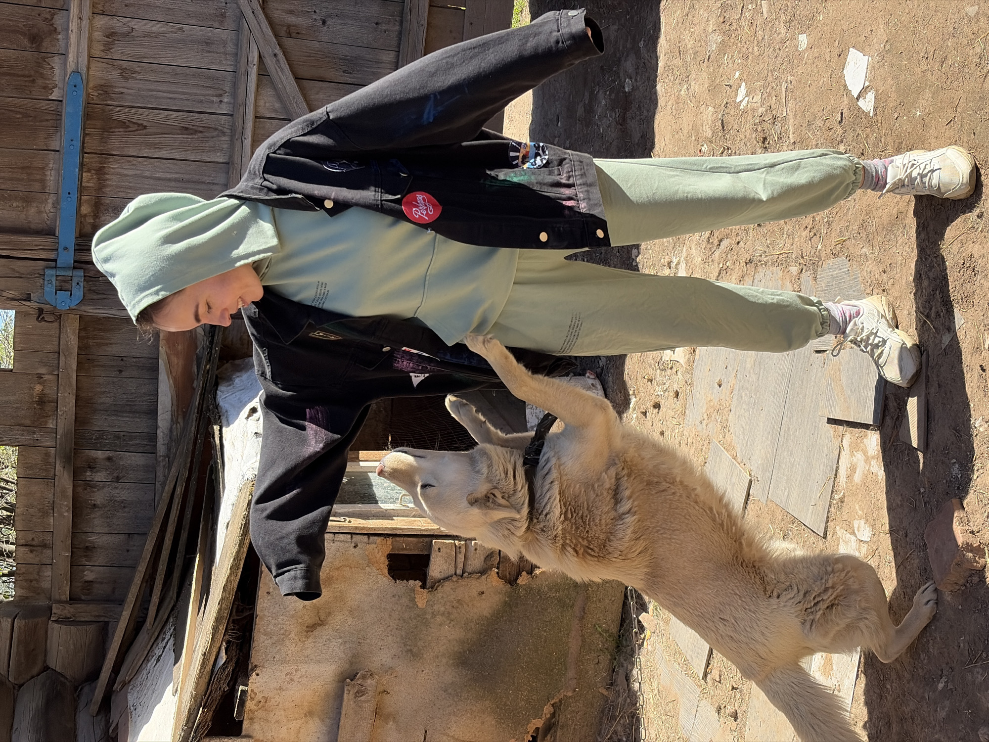 The image depicts a person and a dog in what appears to be a rural or semi-rural setting. The person is wearing a light green hooded sweatshirt, matching sweatpants, a black jacket with patches, and white sneakers. They are standing outside a weathered wooden structure, possibly a shed or an old building. A light-colored dog is standing on its hind legs, seemingly interacting with the person. The ground is a mix of dirt, grass, and scattered pieces of wood. The location is Nizhnii Vishur, Russia.