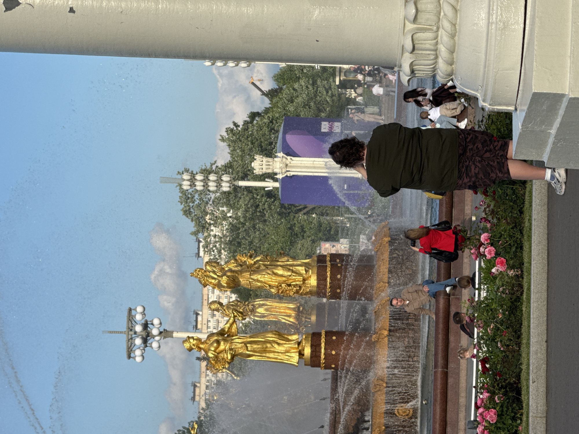 The image depicts the Friendship of Peoples Fountain, a prominent landmark in Moscow, Russia, located at VDNKh Park. The fountain features four golden statues of women representing Soviet republics. In the foreground, a woman in a dark green t-shirt and camouflage skirt stands with her back to the camera, holding her phone up. To her left, a man in a beige jacket and blue jeans sits on the edge of the fountain. To the right of the man, a woman in a red jacket is photographing something with her phone. Several other people are visible in the background, some sitting on the fountain's edge, others standing or walking. The fountain is surrounded by greenery and trees, with a bright blue sky overhead.