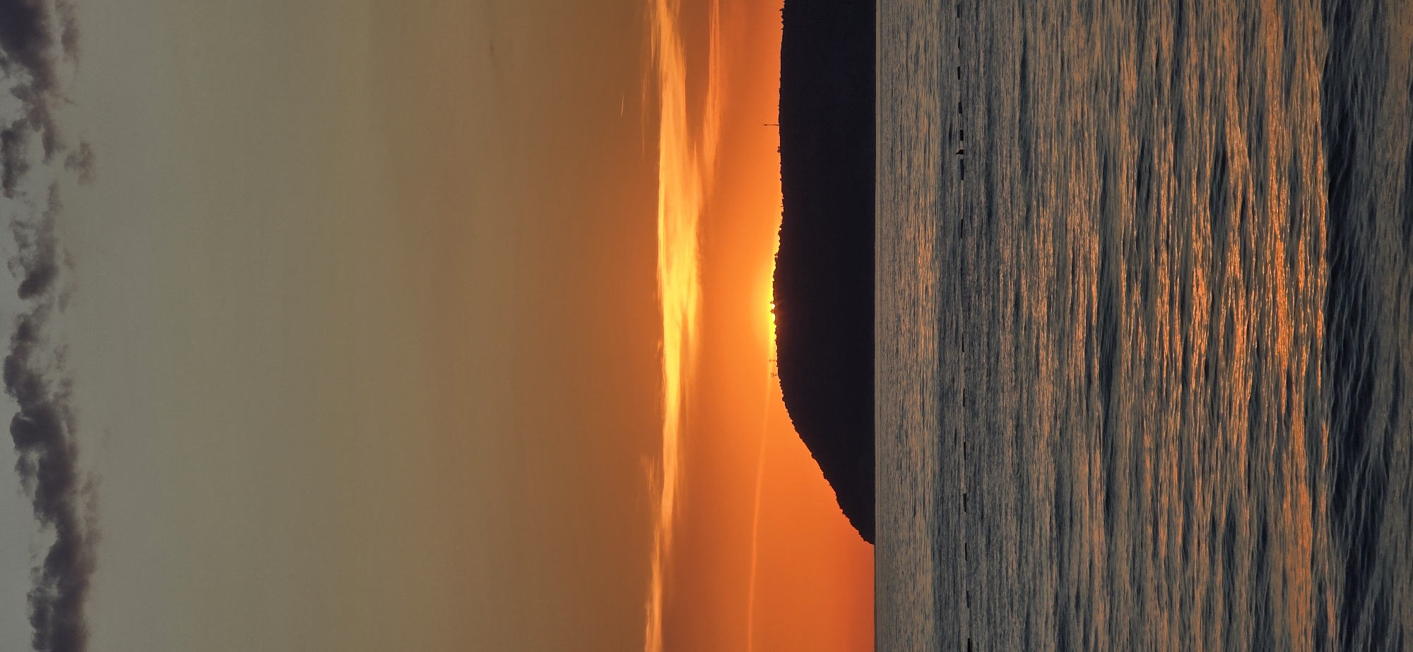 The image shows a landscape at sunset. In the sky, there is an orange glow, suggestive of the setting sun, with some clouds visible on the left side. In the center, there is a dark landmass, possibly a hill or small mountain, that cuts across the orange sky. To the right, the image captures water with reflections of the sunlight, creating a shimmering effect. The overall scene appears to be a sunset over water with land in the background. This scene is consistent with images from the coastal city of Bar, Montenegro.