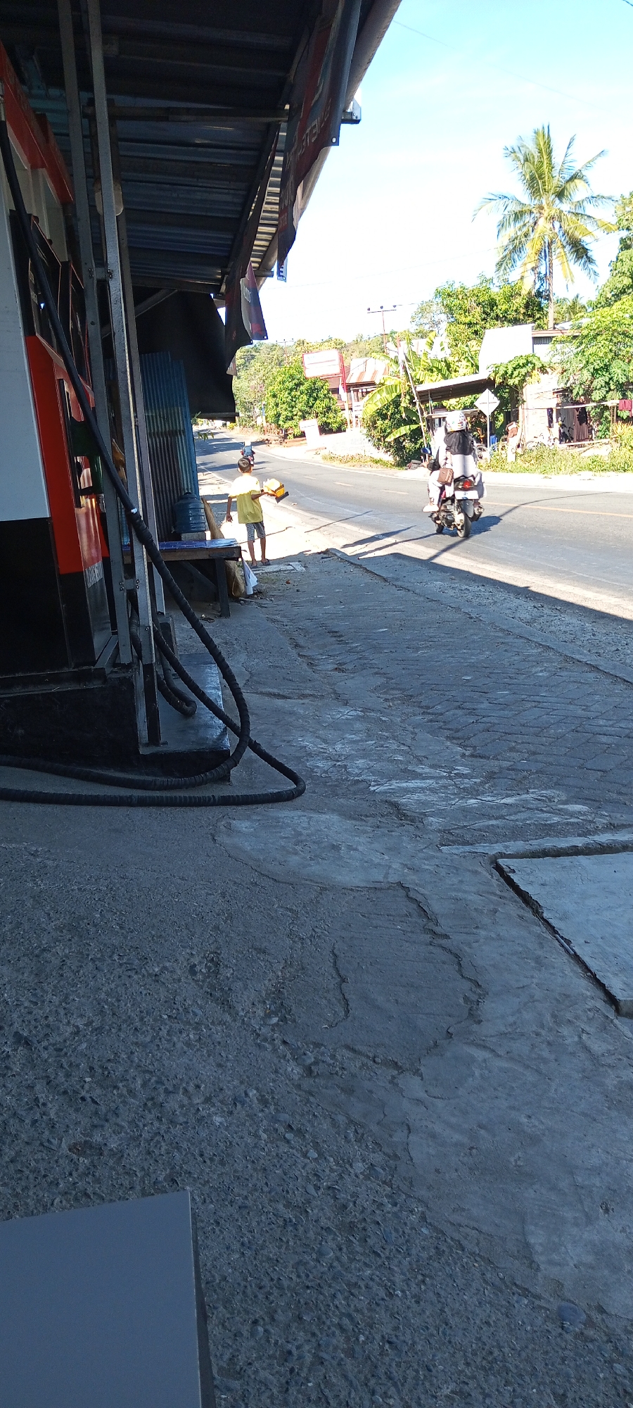 A person stands near a gas pump in Pakkanna, Indonesia, holding a yellow object. A motorcycle with two occupants travels on the road in the background, which is lined with trees and buildings. The scene appears to be a roadside area with a gas station.