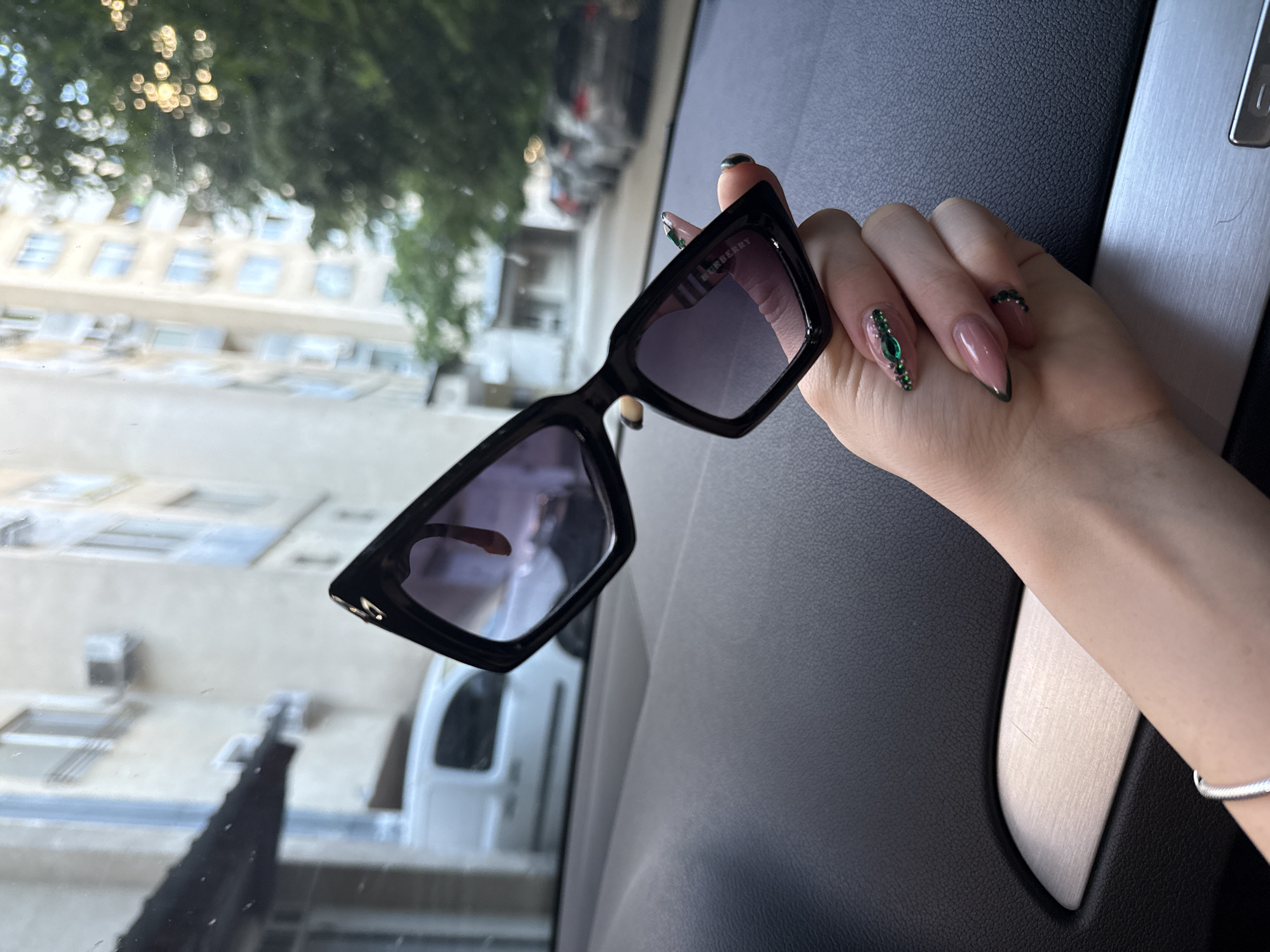 The image shows a woman's hand holding a pair of Burberry sunglasses inside a vehicle. The hand features long, acrylic nails adorned with green jewels and dark tips. The background visible through the car window suggests an urban environment, with buildings and trees. Based on the architecture and foliage visible, the location might be in Plovdiv, Bulgaria, but this is not definitive from the image alone.