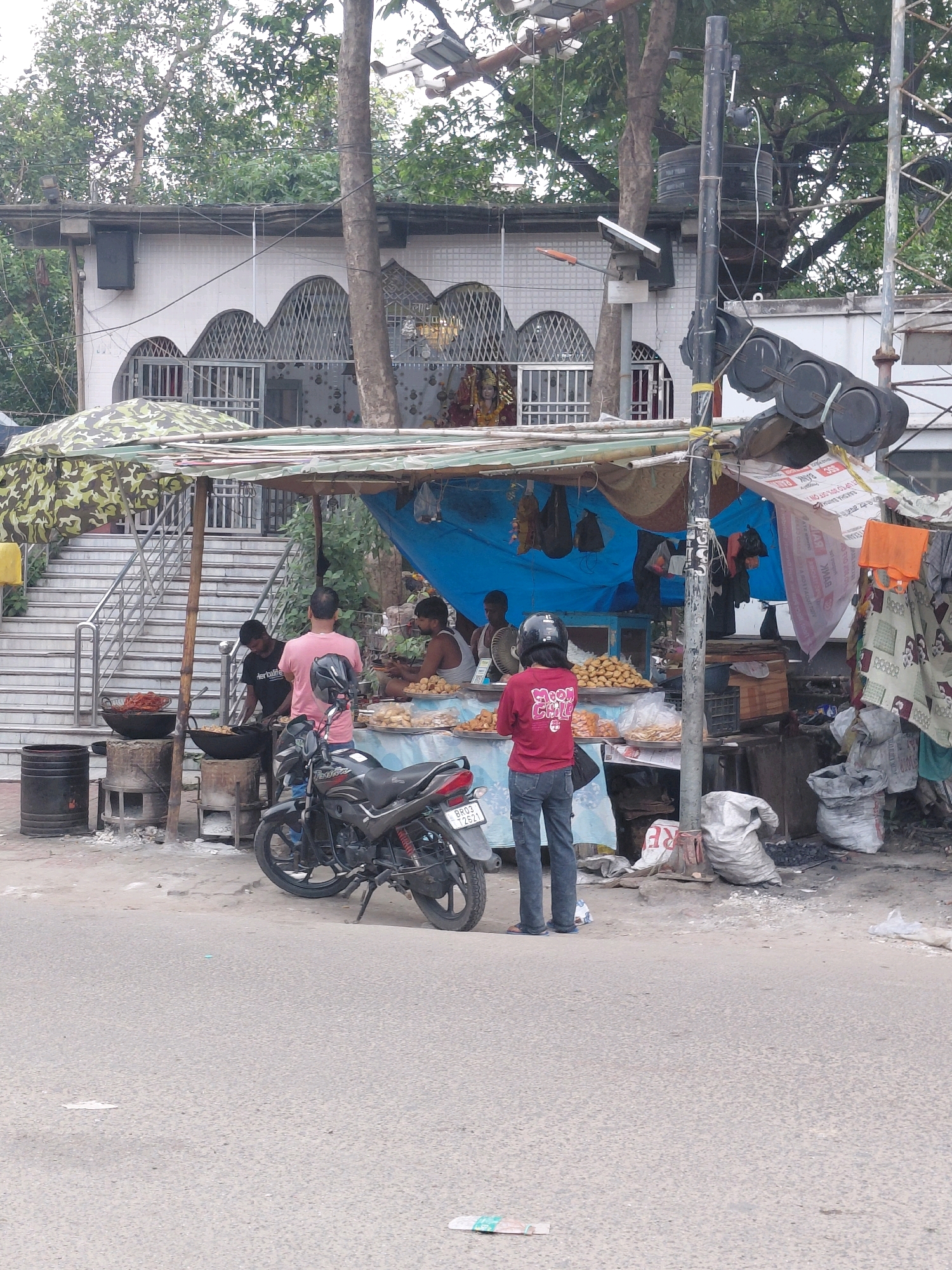 The image shows a street food stall in Patna, India. A motorcycle is parked in front of the stall, which is covered by a camouflage-patterned awning. The stall is selling various fried snacks, including samosas, and appears to be busy with customers and vendors. A man in a pink shirt is standing at the stall, and another man in a black t-shirt is cooking at a separate cooking station to the left. A person wearing a red shirt and a helmet is standing in front of the stall, facing away from the camera. In the background, a building with arched windows and decorative metalwork can be seen, likely a temple or a religious institution. The scene is typical of a bustling street in Patna, with vendors and customers interacting in an outdoor setting. The overall atmosphere suggests a vibrant local market or street food hub.
