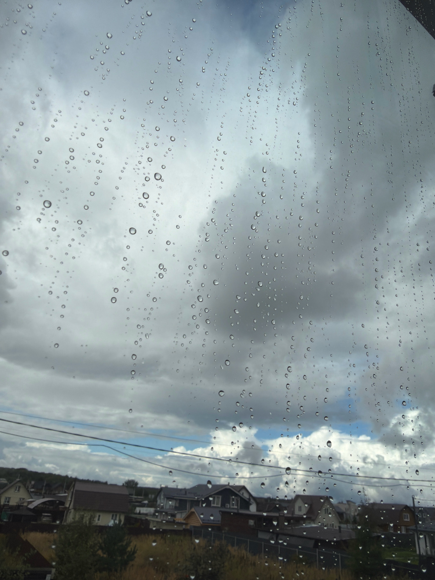 A vertical, slightly low-angle, medium shot captures a cloudy sky filled with raindrops on a windowpane, with a village in the background. The sky is overcast with heavy, gray clouds, and some patches of lighter blue sky are visible. Raindrops, varying in size from small dots to larger spheres, are scattered across the foreground, creating a blurred effect on the glass.
In the background, a village is visible under the cloudy sky. Several houses with dark-colored roofs are clustered together. Power lines run horizontally across the scene, above the houses. The vegetation in the foreground is blurred, appearing as a dark, indistinct mass. The image appears to be taken from inside a building, looking out through a rain-streaked window. The scene suggests a somber and damp atmosphere. There are no people or animals visible in the image. The location is identified as Stupino, Russia.