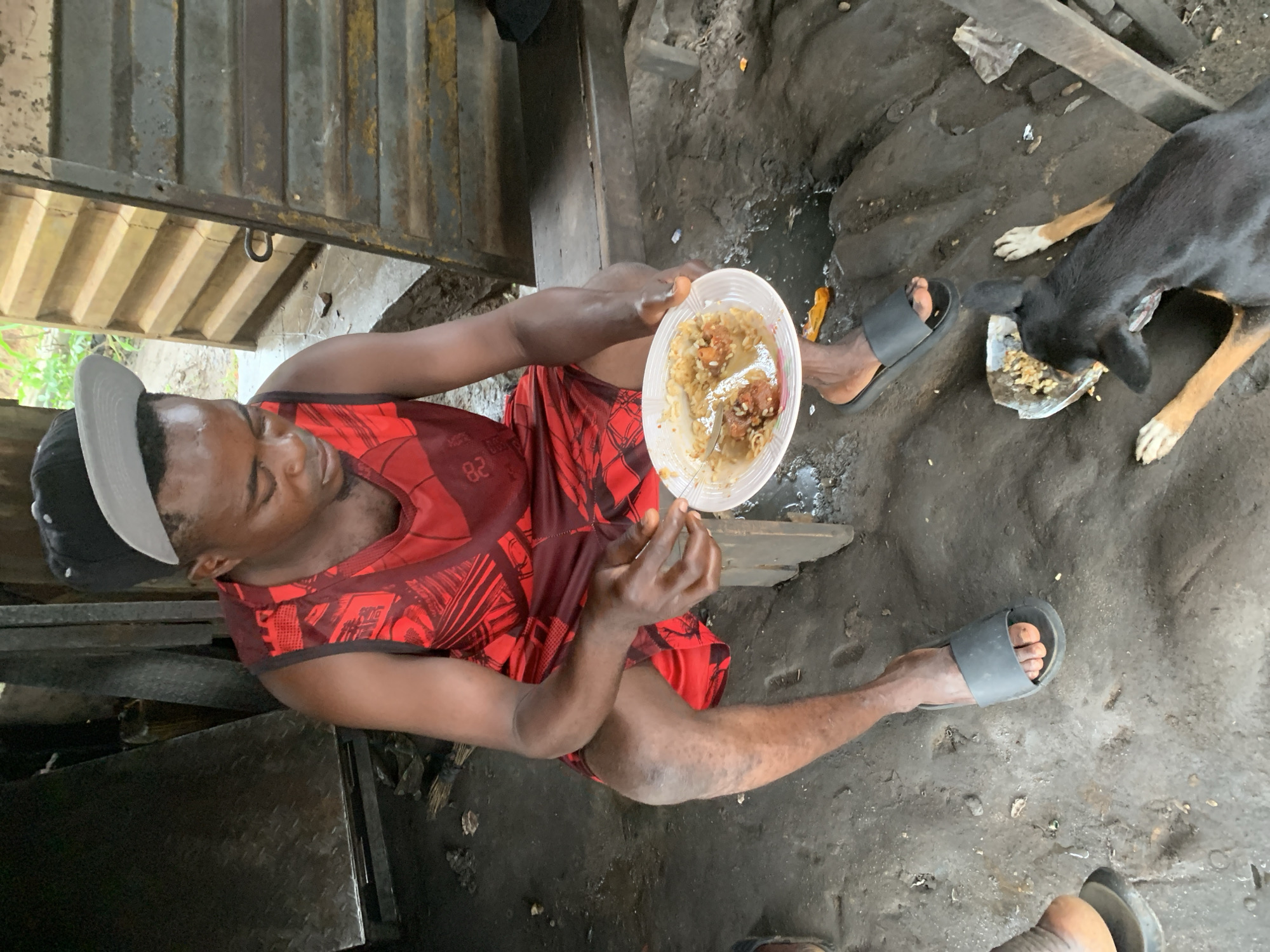 In Shagamu, Nigeria, a Black man wearing a red and black sleeveless shirt and black slides sits on a wooden bench. He is holding a white paper plate filled with rice and meat. A black and tan dog is on the ground, eating from a separate white paper plate. The ground is dirt and littered with debris. In the background, a corrugated metal structure suggests an outdoor market or workshop. The overall scene depicts a simple meal in a rustic, possibly impoverished, urban setting.