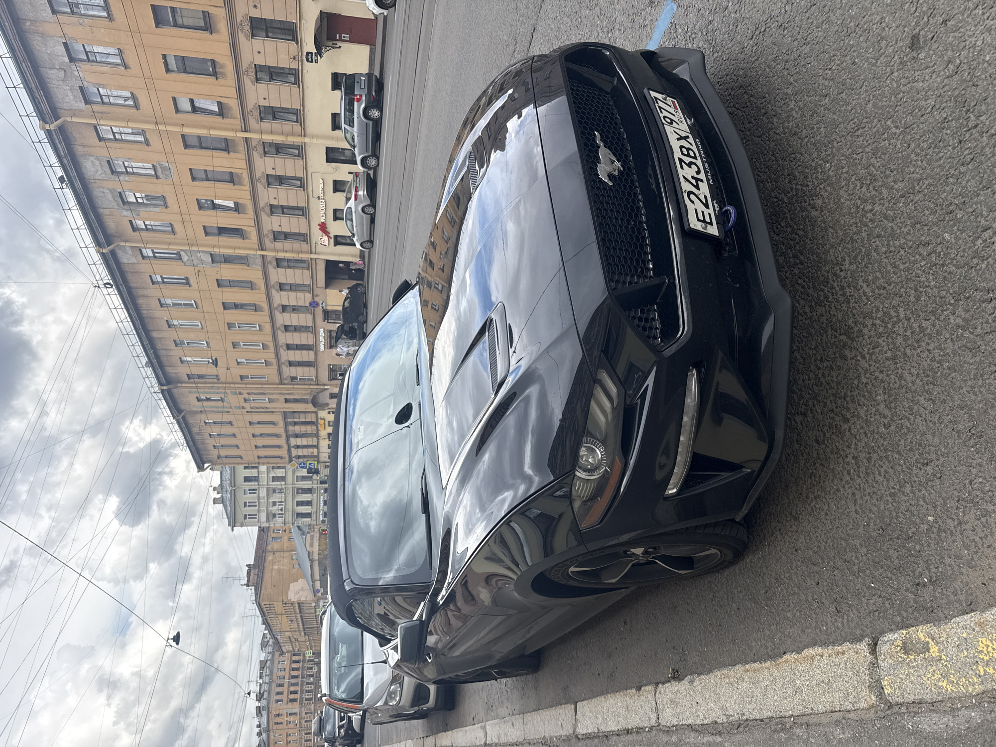 A black Ford Mustang convertible is parked on a city street in Saint Petersburg, Russia, with its front passenger side facing the viewer and angled slightly towards the road. The car is parked along a sidewalk with a yellow and grey curb. The street is lined with multi-story buildings, characteristic of Saint Petersburg's architectural style, with yellow and tan facades and numerous windows. Other vehicles, including a silver sedan and a silver SUV, are visible parked further down the street. Overhead, a complex network of wires crisscrosses the sky, common in urban environments. The sky is overcast with clouds. The Mustang's license plate is visible and reads "E243BX 977 RUSSIA". A blue marking is visible on the asphalt in front of the car.