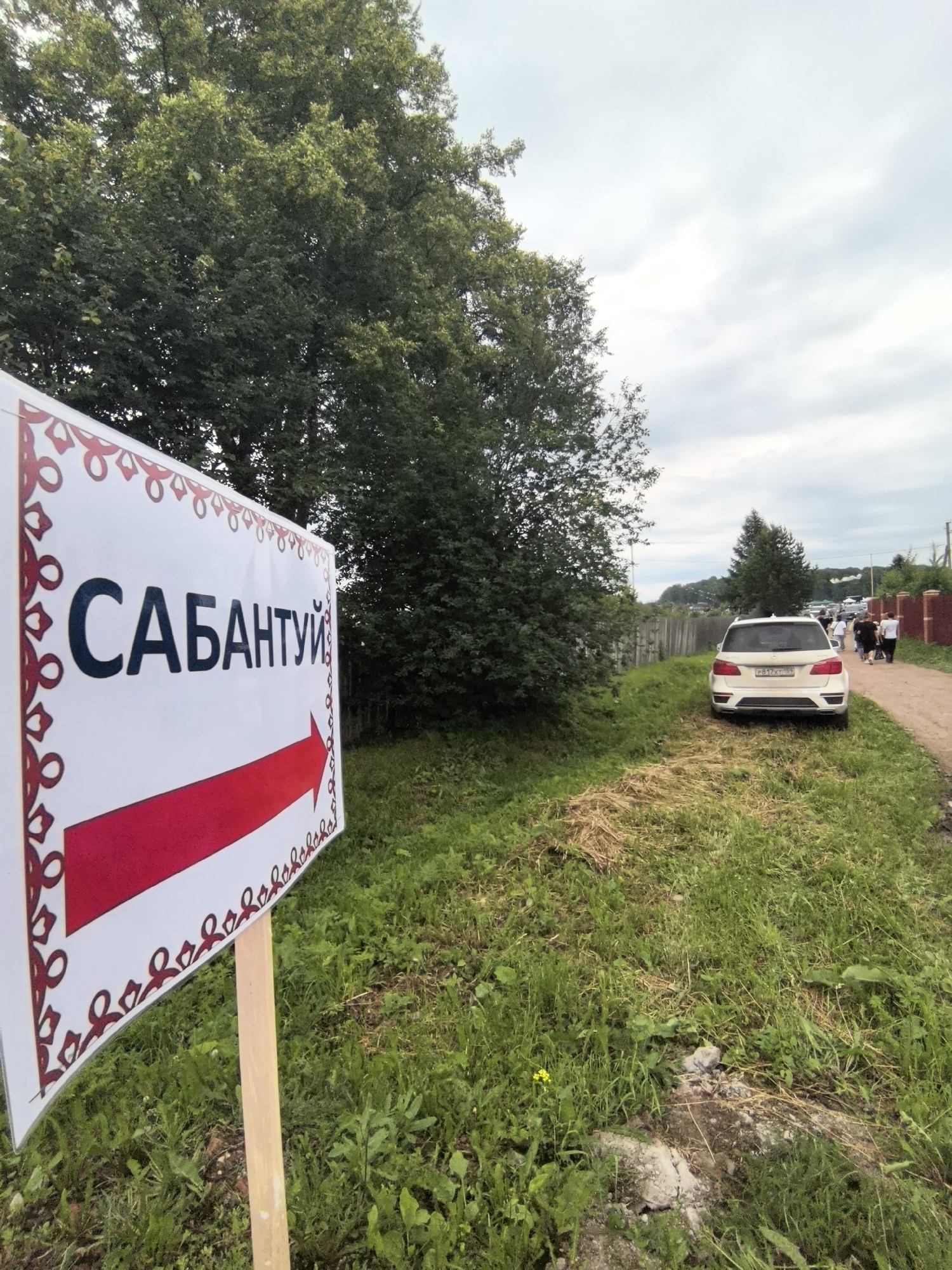 A white sign with a red arrow pointing to the right and the Cyrillic word "САБАНТУЙ" is planted in the grassy area next to a dirt road. The sign has decorative red trim on its left side. In the background, several people are walking down the road past a white SUV. The scene appears to be in a rural or suburban area with trees and houses visible in the distance. The location is identified as Кояново, Russia.
