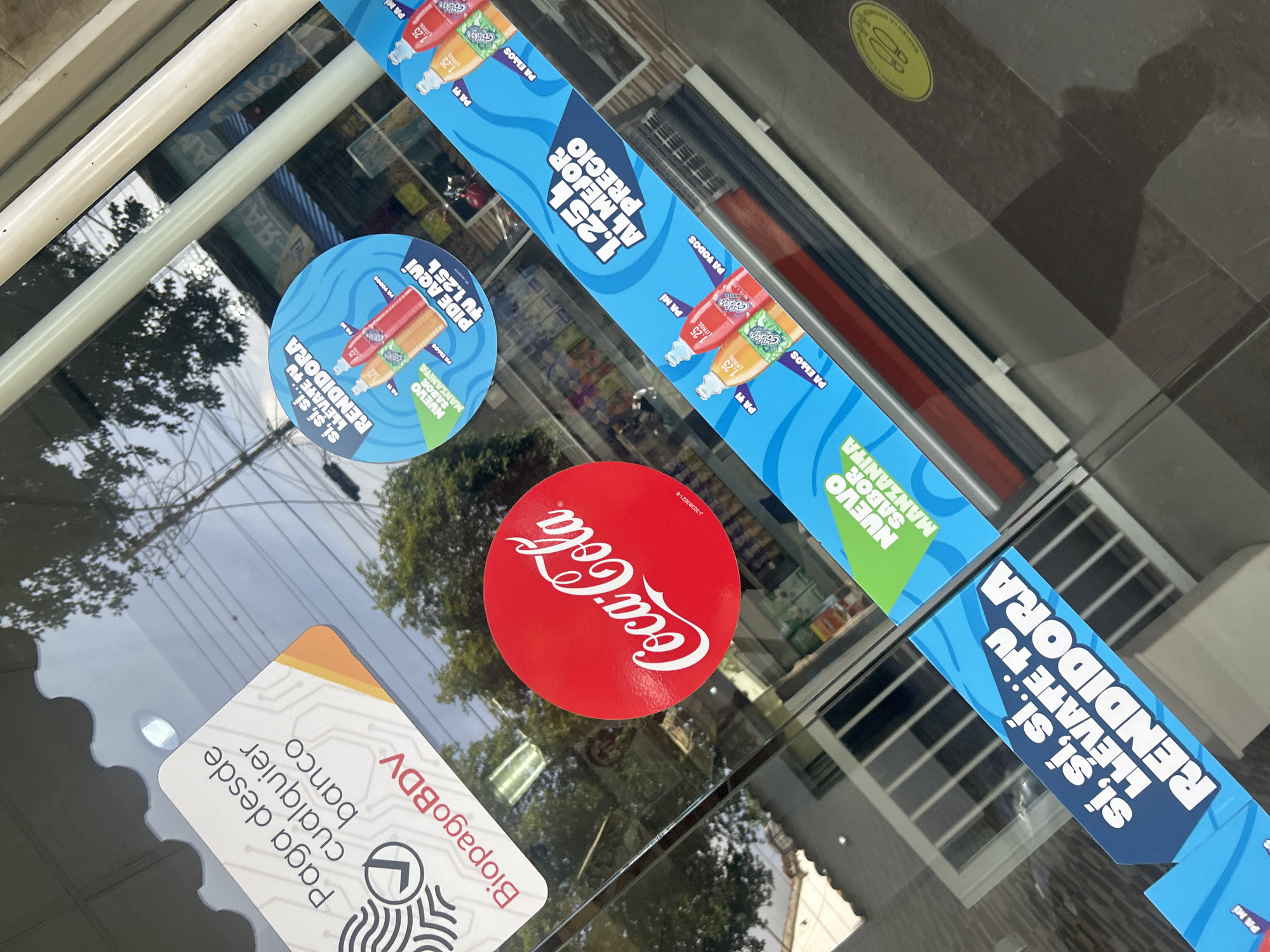 In the city of Santa Ana, Venezuela, a glass storefront displays various stickers and signage. On the left, a white and orange sticker reads "Paga desde cualquier banco" and "BiopagoBDV." To its right, a prominent red circular sticker features the "Coca-Cola" logo. Further to the right, a blue diagonal sticker strip showcases images of Coca-Cola bottles and text like "Sí, sí, sí... LLEVATE TU RENDIDORA" and "NUEVO SABOR MANZANITA." Another blue sticker strip below also advertises "1.25 L AL MEJOR PRECIO" with bottle images. The reflection on the glass shows trees, power lines, and building structures. The floor inside the store appears to be tiled with dark brown and lighter brown squares, and a yellow circular sticker is visible on the floor towards the bottom right. The overall scene captures commercial advertising on a store entrance.