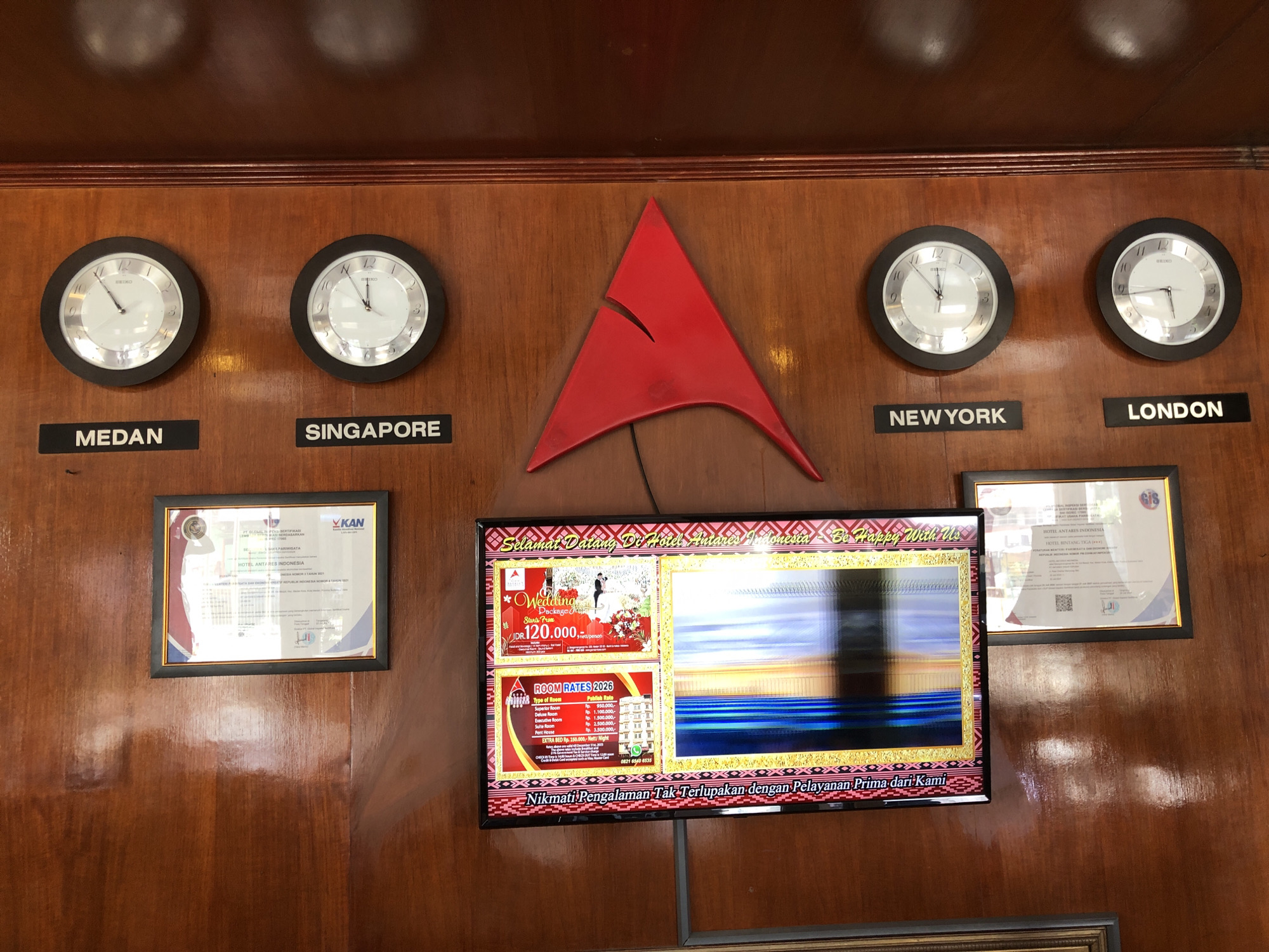 The image shows a wall with a wooden panel design. Four clocks labeled Medan, Singapore, New York, and London show the respective times. A red triangular logo is positioned in the center of the wall. Beneath the logo is a television screen displaying a hotel advertisement and information regarding wedding packages and room rates for the Hotel Antares Indonesia. The screen also displays a phone number. Two framed certificates are located on either side of the television. The bottom text on the TV translates to "Enjoy an Unforgettable Experience with First Class Service from Us". The OCR text matches the text on the screen.