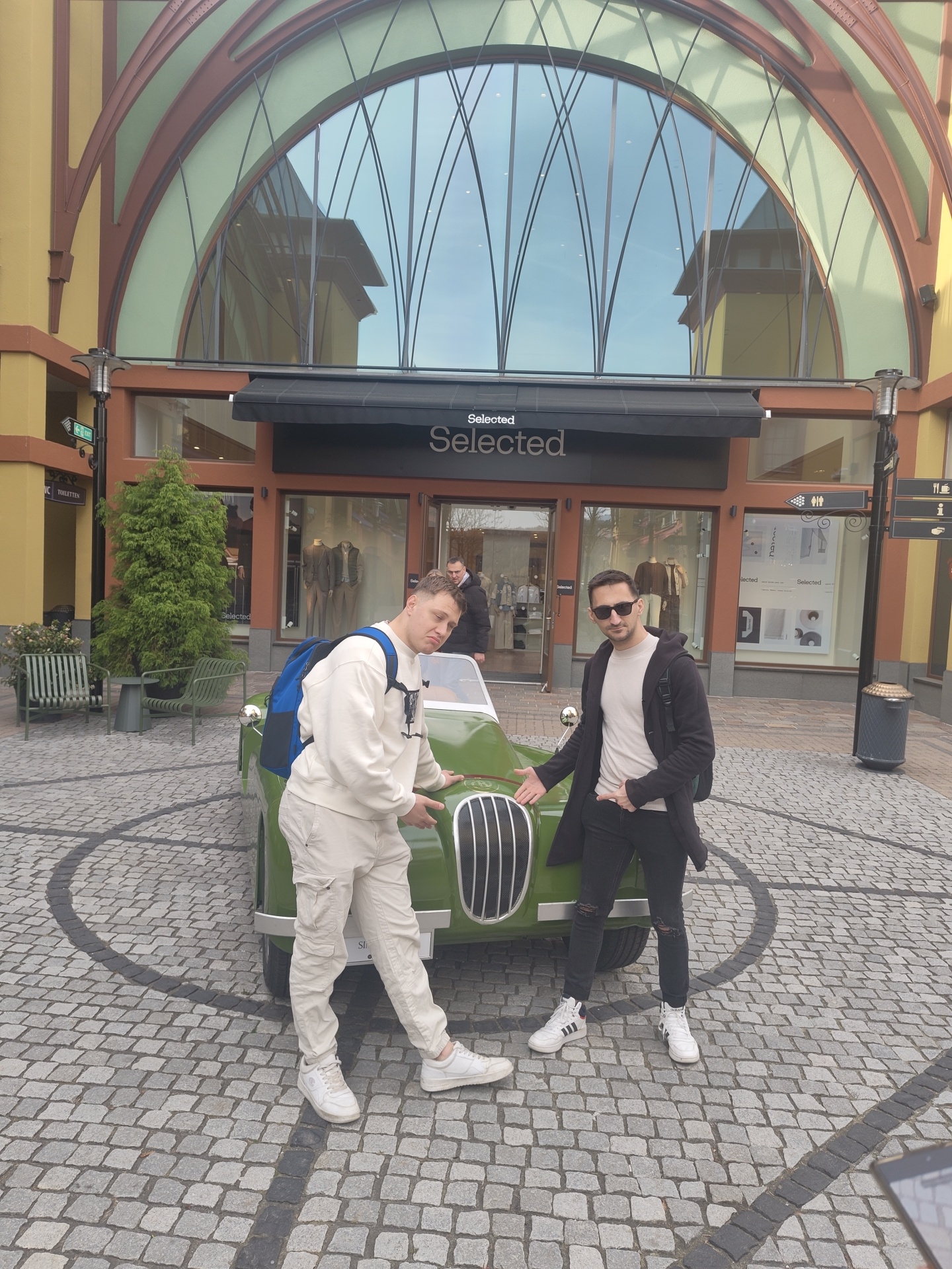 Two adult males are posing with a small, olive green, vintage-style roadster automobile in an outdoor, cobblestone plaza at Wertheim Village, Wertheim, Germany.
The male on the left wears a cream-colored sweatshirt, matching cargo pants, and white sneakers, with a blue backpack on his back. He leans on the car's hood, looking towards the camera with a slight expression. The male on the right wears sunglasses, a light t-shirt, a black long coat, black ripped jeans, and white high-top sneakers. He stands beside the car's front fender, gesturing with his right hand towards the car while looking directly at the camera.
The car, featuring a chrome grille and bumper, is positioned on a cobblestone surface marked with a distinctive black circular pattern.
In the background, a multi-story building houses a "Selected" clothing store at ground level, identifiable by its black awning and storefront signage. The building facade is primarily yellow with brown trim. Above the store, a large arched window with a complex, curved metal framework is prominent, reflecting parts of the surrounding architecture and sky. To the left of the store entrance are a small green evergreen tree and several green metal outdoor chairs and a bench. To the right, a black trash receptacle is visible. Signs on poles indicate "Toiletten" (restrooms) and directional arrows for other amenities. A third male is partially visible inside the "Selected" store, looking towards the subjects.
