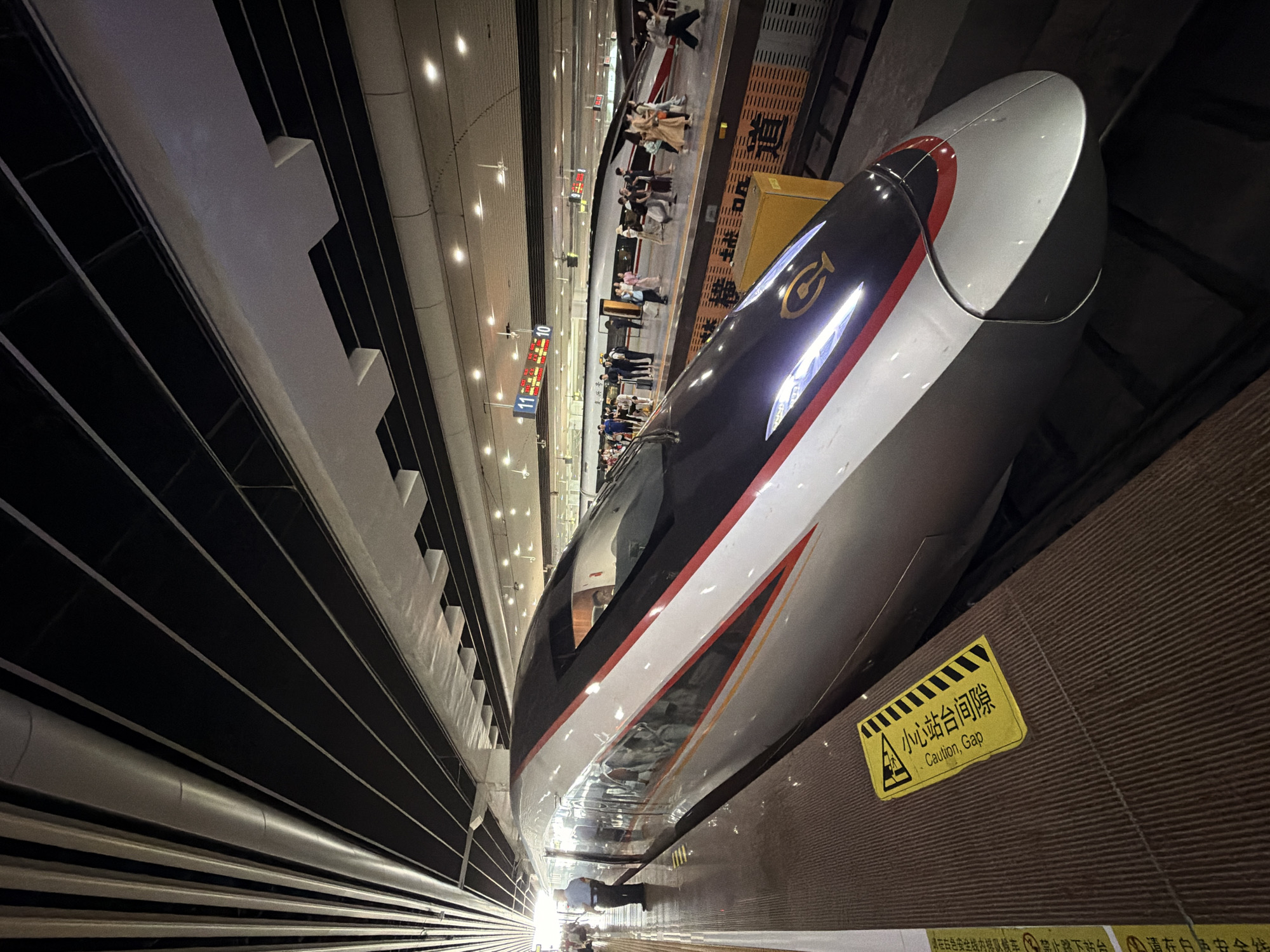 A high-speed train with a silver and red design is at a train station platform in Panyu District, China. The train's headlights are illuminated. A caution gap sign is visible on the platform next to the train. People are standing on the platform, some with luggage. The station has a modern ceiling structure with exposed beams and lighting. In the background, other trains and platforms are visible, along with digital display boards showing train information. The lighting suggests it is daytime or a well-lit indoor environment.
