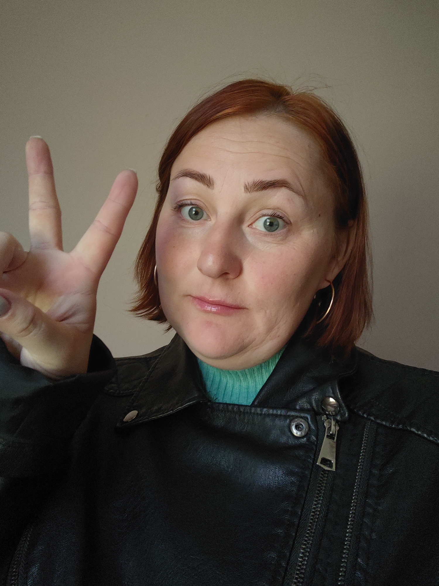 A woman with fair skin, red hair, and green eyes is making a three-finger gesture. She has a bruise under her left eye. She is wearing a black leather jacket over a turquoise turtleneck sweater and gold hoop earrings. The background is a solid light beige color. The location is not evident from the image.