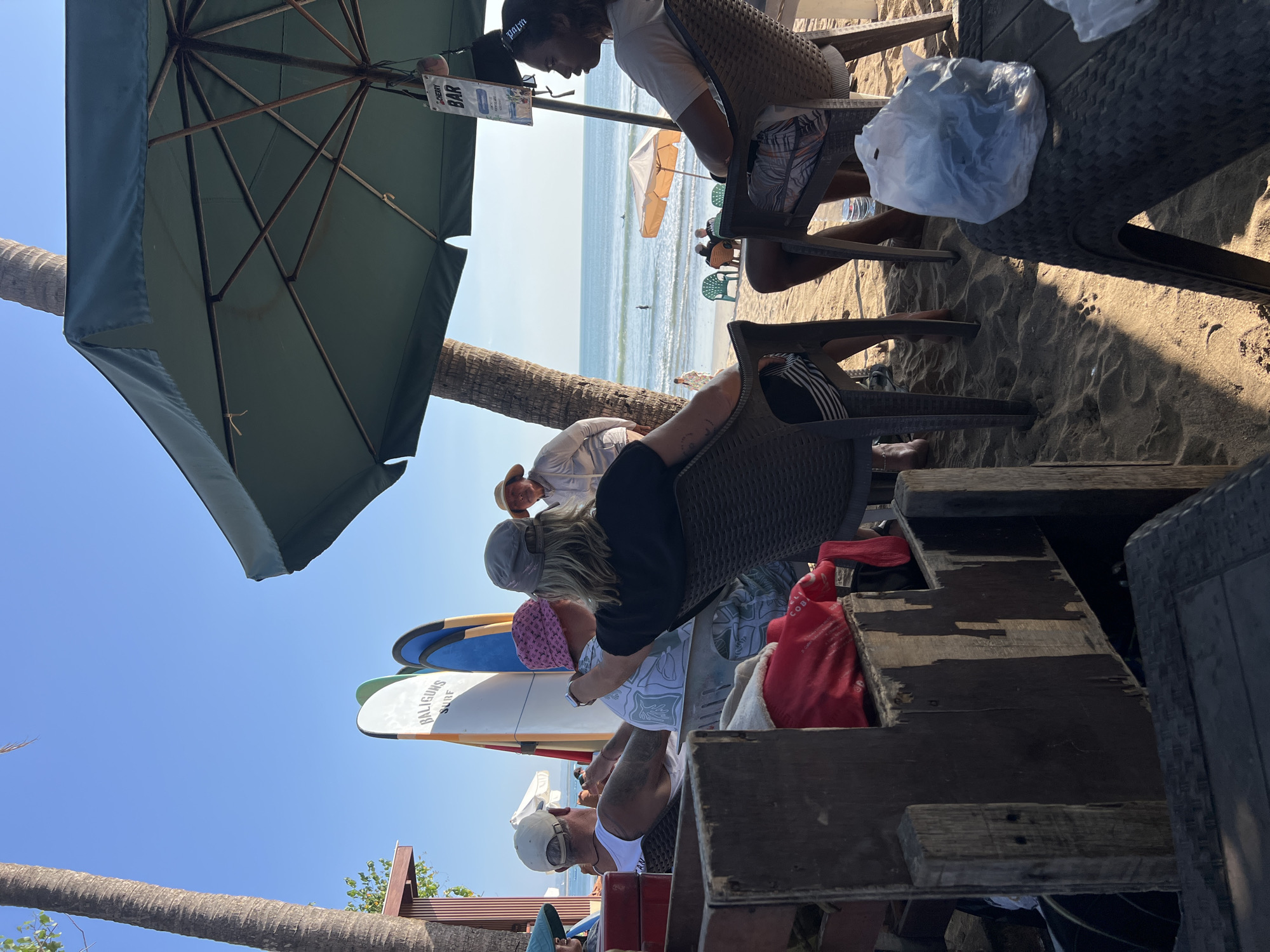 The image depicts a beach scene in Kuta, Indonesia, on a clear day with the ocean in the background. Several people are seated under a large green umbrella, seemingly at a beachside bar or cafe. In the foreground, there are wooden structures and tables with plastic bags. Two surfboards with the inscription "BALIGUNS SURF" are propped up behind the seated individuals. The people are casually dressed, with some wearing hats. The atmosphere appears relaxed, with some individuals looking towards the ocean where a few people are in the water and others are walking along the shore. The sand is golden, and palm trees are visible. A sign attached to the umbrella pole reads "CHERRY BAR."