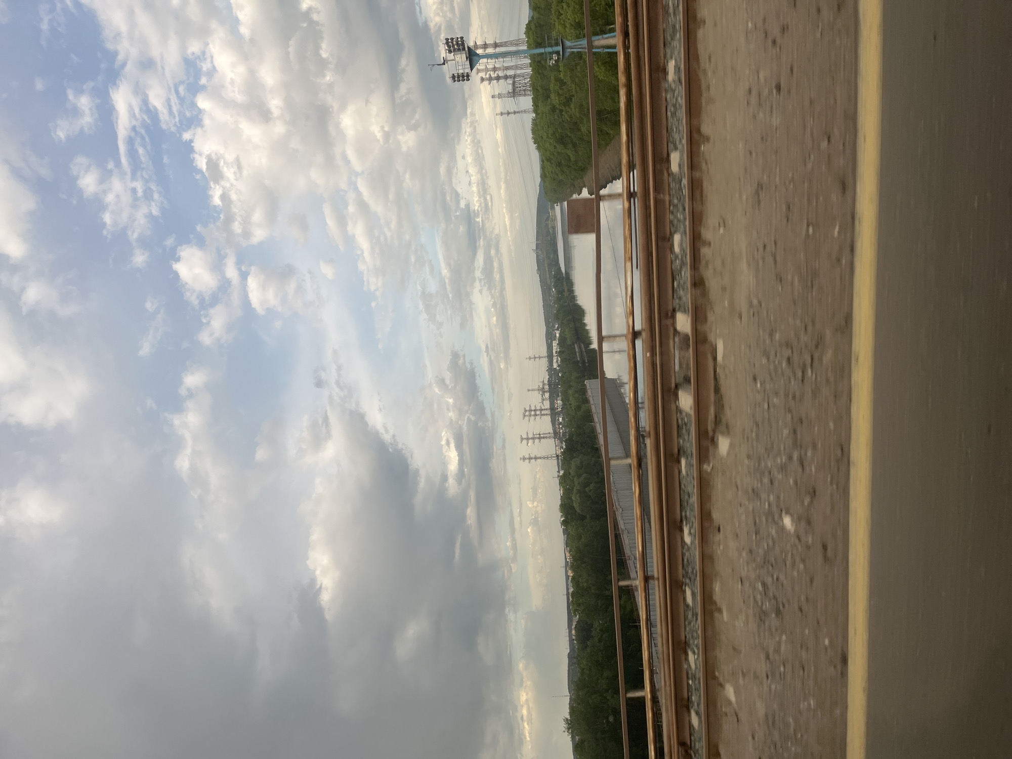 The image presents a view from a bridge or elevated structure. The sky is filled with various cloud formations, ranging from light and wispy to darker, more dense clusters. Below, there's a waterway, possibly a river or a wide canal, stretching into the distance, reflecting the sky and light. The banks of the waterway are lined with greenery, including trees and vegetation. In the distance, there are structures and utility poles, suggesting an urban or industrial area. The presence of high-voltage power lines indicates the proximity of industrial or power-generating facilities. The foreground features metal railings and a concrete surface. The yellow line on the side could indicate that the viewer is on a highway bridge. The location is Chaikovsky, Russia, based on the high-voltage power lines and typical landscapes of the Kama River in the Chaikovsky area. There are no people directly visible in the frame.