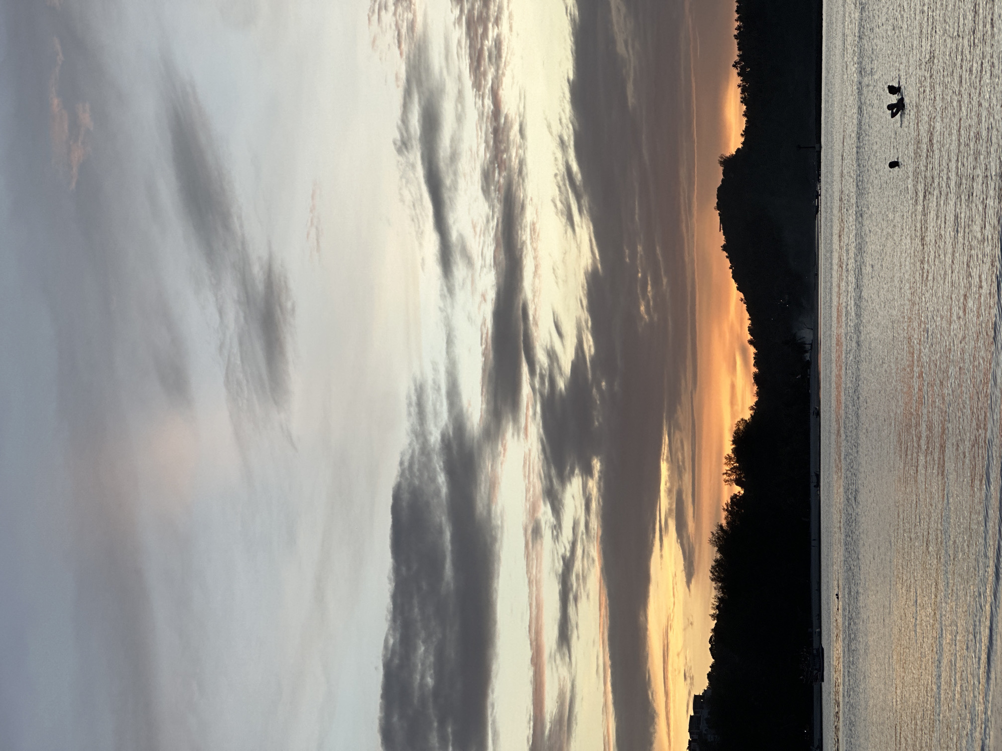 The image depicts a serene coastal sunset scene at Ao Phai, Koh Phangan, Thailand. The sky is a gradient of soft grays and pale oranges, with wispy clouds streaking across, catching the last rays of the setting sun. The water in the foreground is calm, reflecting the muted colors of the sky. Silhouetted against the horizon are lush green trees and the dark outlines of hills, with a few structures visible on the left side of the frame. In the water, a small group of people, their figures dark against the light, are either swimming or wading. A small boat is also visible in the distance near the shore. The overall impression is one of tranquility and the natural beauty of a tropical evening.