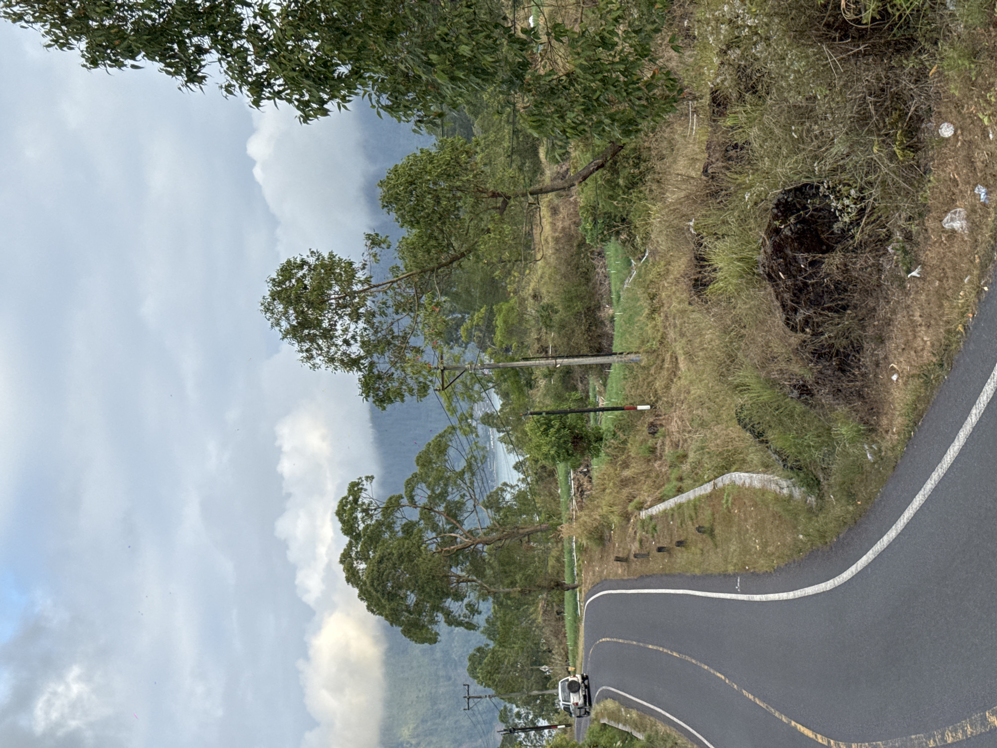 A white SUV drives along a winding paved road in Kintamani, Indonesia. The road curves to the left, with yellow and white lane markings. On the right side of the road, dry grass and bushes grow alongside a low concrete barrier. A few wooden posts are visible along the edge of the road. In the background, there are several trees with lush green foliage, and further in the distance, a body of water is visible, likely a lake, surrounded by misty mountains. The sky is overcast with clouds. A few utility poles with wires are also present along the road. The overall scene depicts a scenic rural landscape with a clear road and natural surroundings.