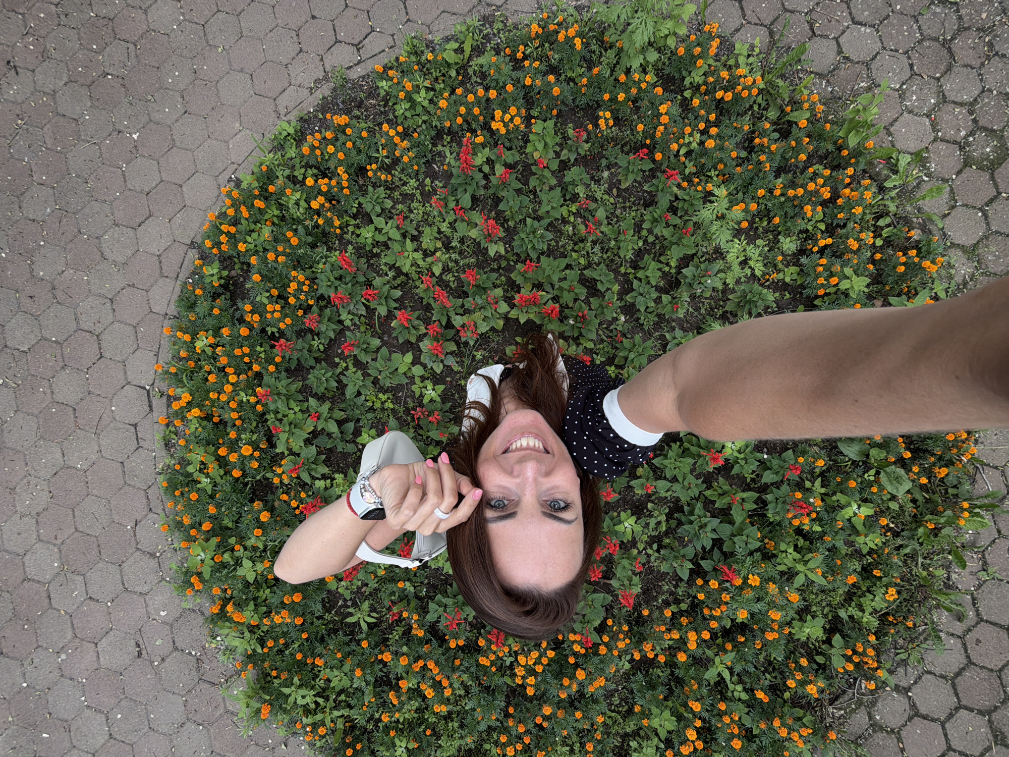 The image is a high-angle, close-up shot of a woman standing in a circular flower bed filled with small orange marigolds and red salvia flowers. The flower bed is surrounded by hexagonal paving stones. The woman, with long brown hair and blue eyes, is smiling at the camera. She is wearing a black and white polka dot dress, a white purse slung over her shoulder, and a watch and ring on her left hand. The arm of the person taking the photo is visible in the foreground. The scene appears to be an outdoor public space within the city of Birobidzhan, Russia, possibly a park or street corner.
