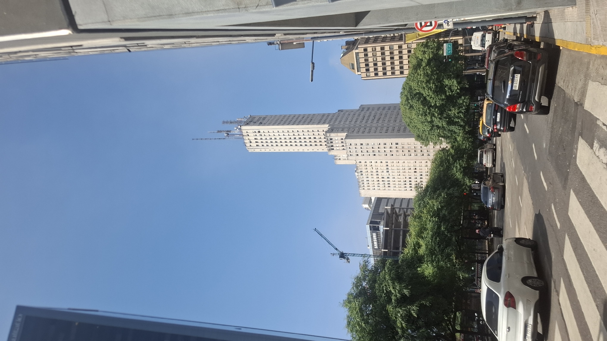 Here is a description of the image:
The image captures a street scene in Buenos Aires, Argentina, featuring a tall, light-colored skyscraper in the background and typical urban elements. The building is rectangular with numerous windows and antennae or construction equipment at the top. The clear blue sky dominates the upper portion of the image.
On the right side of the image, a street with cars parked and in motion is visible. Crosswalks mark pedestrian paths. Trees line the street, partially obscuring the view of the buildings behind them. Other buildings of varying heights and architectural styles are visible adjacent to the main skyscraper.
The image is taken from a slightly elevated perspective. There are no visible people. The environment suggests a busy urban area during daylight hours.