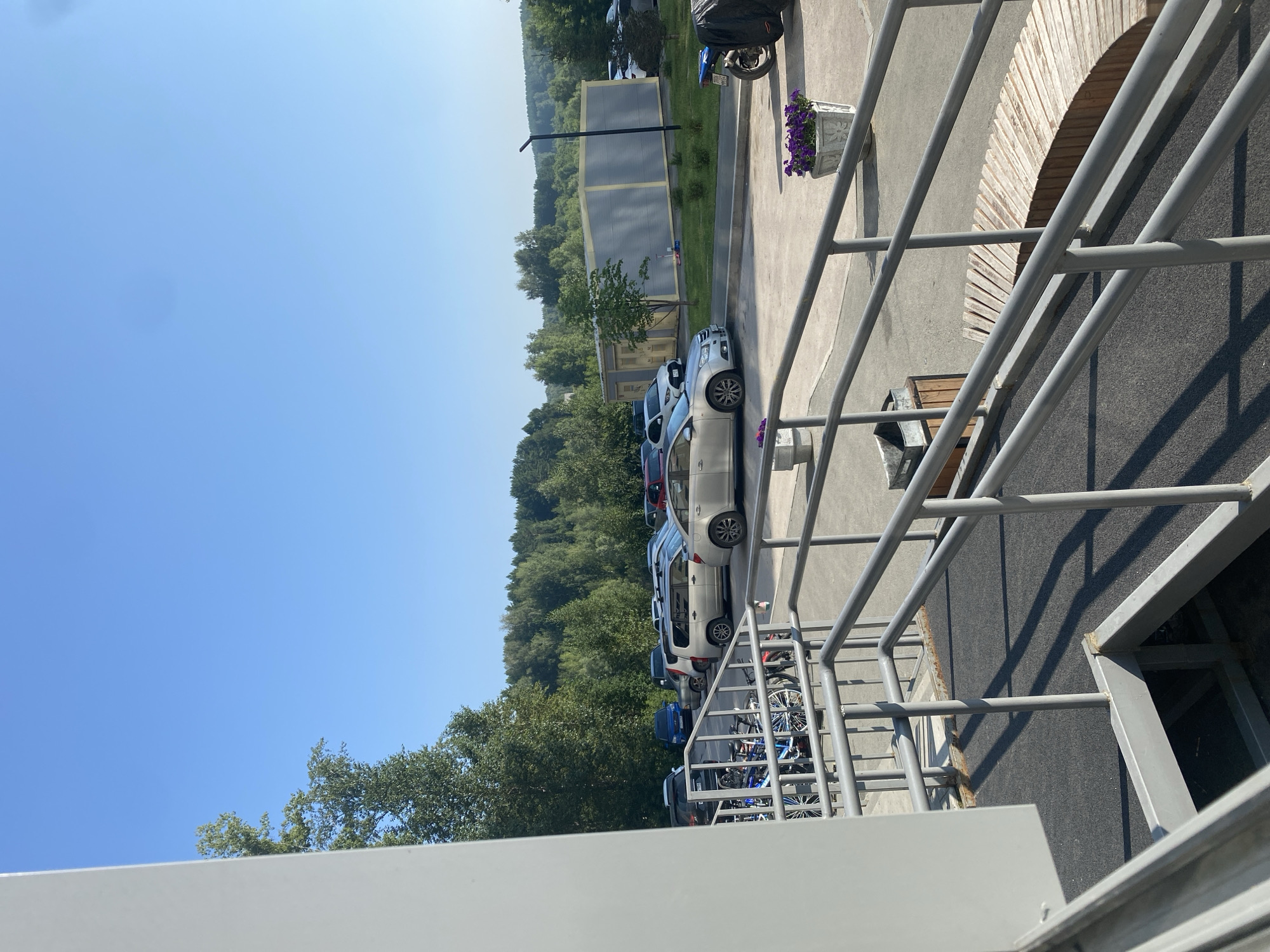 The image captures an outdoor scene on a sunny day, likely in Novosibirsk, Russia, given the context. A grey, metal railing system, part of a ramp or elevated walkway, dominates the foreground. Beyond the railing, a parking lot is visible, filled with various cars including a silver sedan, a silver minivan, and a dark blue SUV. In the middle ground, a row of trees forms a dense green backdrop, with a long, grey and yellow modular building situated behind the parking lot. The sky is clear and blue. To the right, a blue motorcycle covered with a black tarp is parked. Two white planters filled with purple flowers are placed on the pavement. In the foreground, shadows are cast on a dark grey asphalt surface.