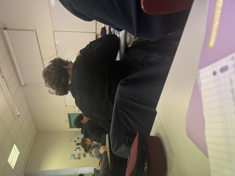 The image depicts a classroom in Albi, France, observed from a low angle, looking towards the front of the room. In the immediate foreground, a light-colored desk surface is visible, bearing a dark red pencil case on the left and a partially visible ruled notebook atop a purple folder on the right.
The central focus is a male individual seated at a desk, seen from behind. He possesses dark, curly hair and wears a black long-sleeved shirt. A dark blue jacket is draped over the back of his chair. He appears to be looking down at his desk. To his left, another male student, also viewed from behind, wears a dark hoodie. Further left and slightly behind the hooded student, a female individual with brown hair, tied back, wears a grey hooded top and is seated at a desk, looking downward. A segment of another individual's arm is visible on the far left. On the far right, the dark blue back and part of a red chair are visible.
The classroom environment includes light-colored walls. The left wall displays multiple papers or posters affixed to it. A light-colored door, possibly teal or light green, is situated on the left wall, near the corner of the front wall. The front wall features two whiteboards and a retractable projection screen positioned above them. A circular clock with a white face is mounted on the front wall between the door and the whiteboards. Overhead, rectangular fluorescent light fixtures are visible on the ceiling. All visible individuals are seated at individual desks.