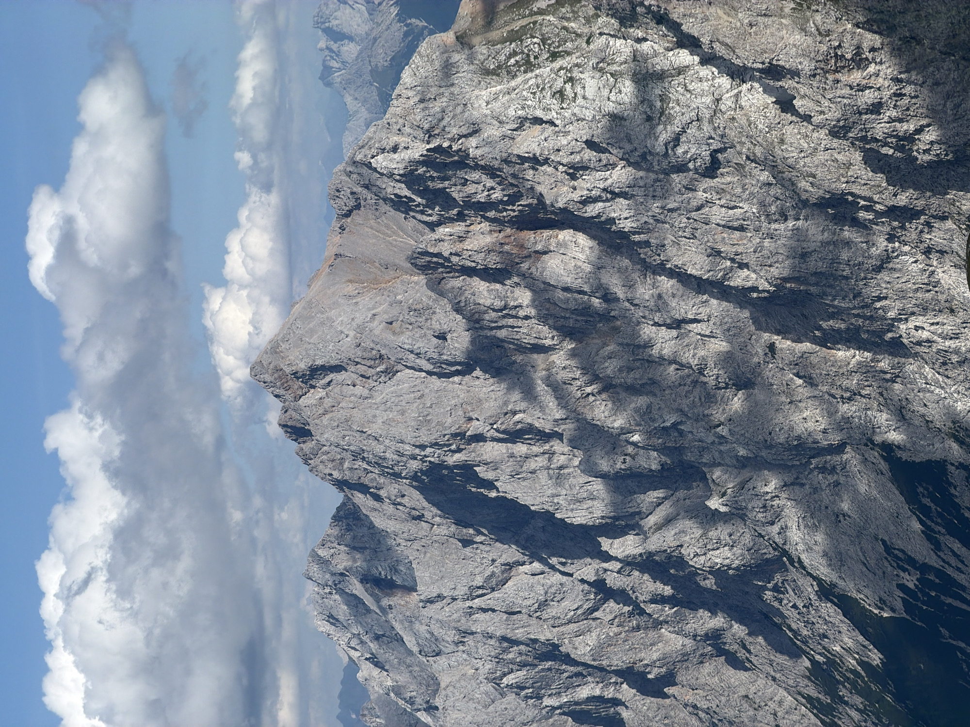 The image is a close-up, aerial shot of a rugged mountain range under a partly cloudy sky. The mountains are predominantly grey, with sharp peaks and deep shadows cast by the clouds. Some patches of green vegetation are visible on the lower slopes. The sky is blue with scattered white cumulus clouds. The scene depicts a natural, mountainous environment, typical of alpine regions. This view is consistent with the landscape surrounding Garmisch-Partenkirchen, Germany, which is renowned for its dramatic mountain scenery, including peaks like the Zugspitze. There are no people or man-made objects visible in the image.