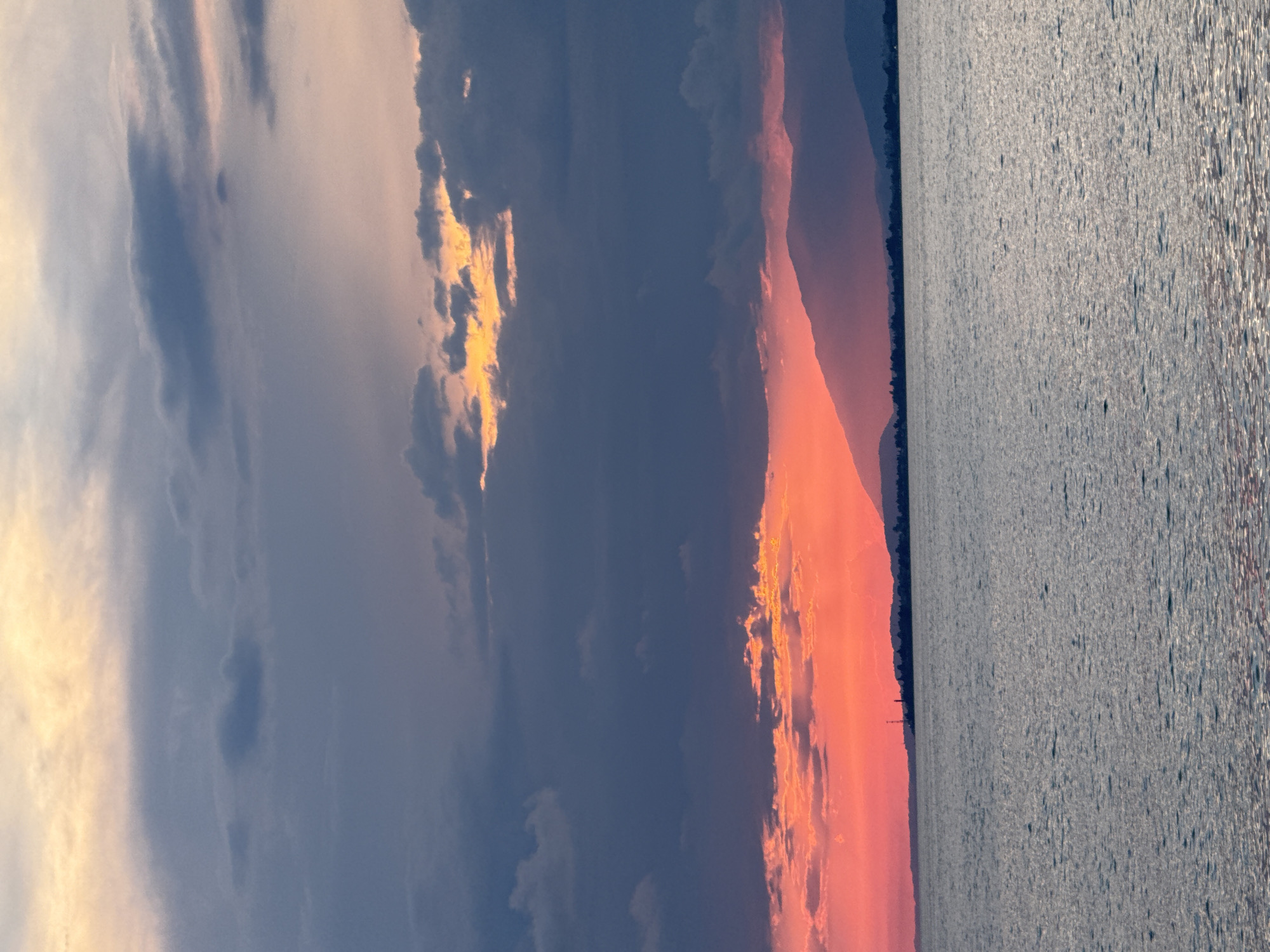 The image captures a serene sunset over the Black Sea near Yuada-Gulrypsh, Abkhazia. The sky is a dramatic canvas of color, transitioning from a fiery orange and pink hue near the horizon, where the sun is setting, to cooler shades of purple and grey clouds higher up. The water in the foreground is calm, reflecting the muted colors of the sky. A silhouette of a coastline with distant hills and a few structures, possibly including a tower, can be seen in the middle ground. The overall scene is tranquil and picturesque, characteristic of a coastal sunset.