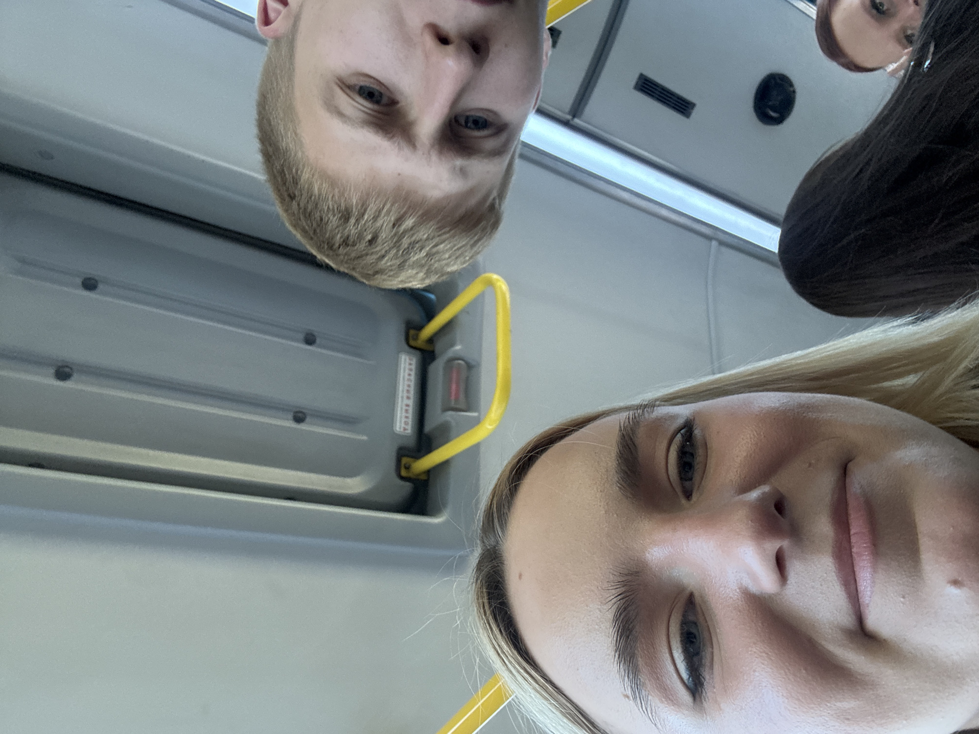The image shows a low-angle selfie taken on what appears to be public transportation, likely a bus or train. Two individuals are prominently featured: a blonde-haired person on the bottom and a lighter-haired person with shorter hair above. The blonde person is partially smiling. A third person is partially visible in the upper right corner. Elements of the interior, such as a yellow grab bar, a gray wall panel, and overhead vents, are visible. The location is unknown and cannot be determined to be Vsevolozhsk, Russia.