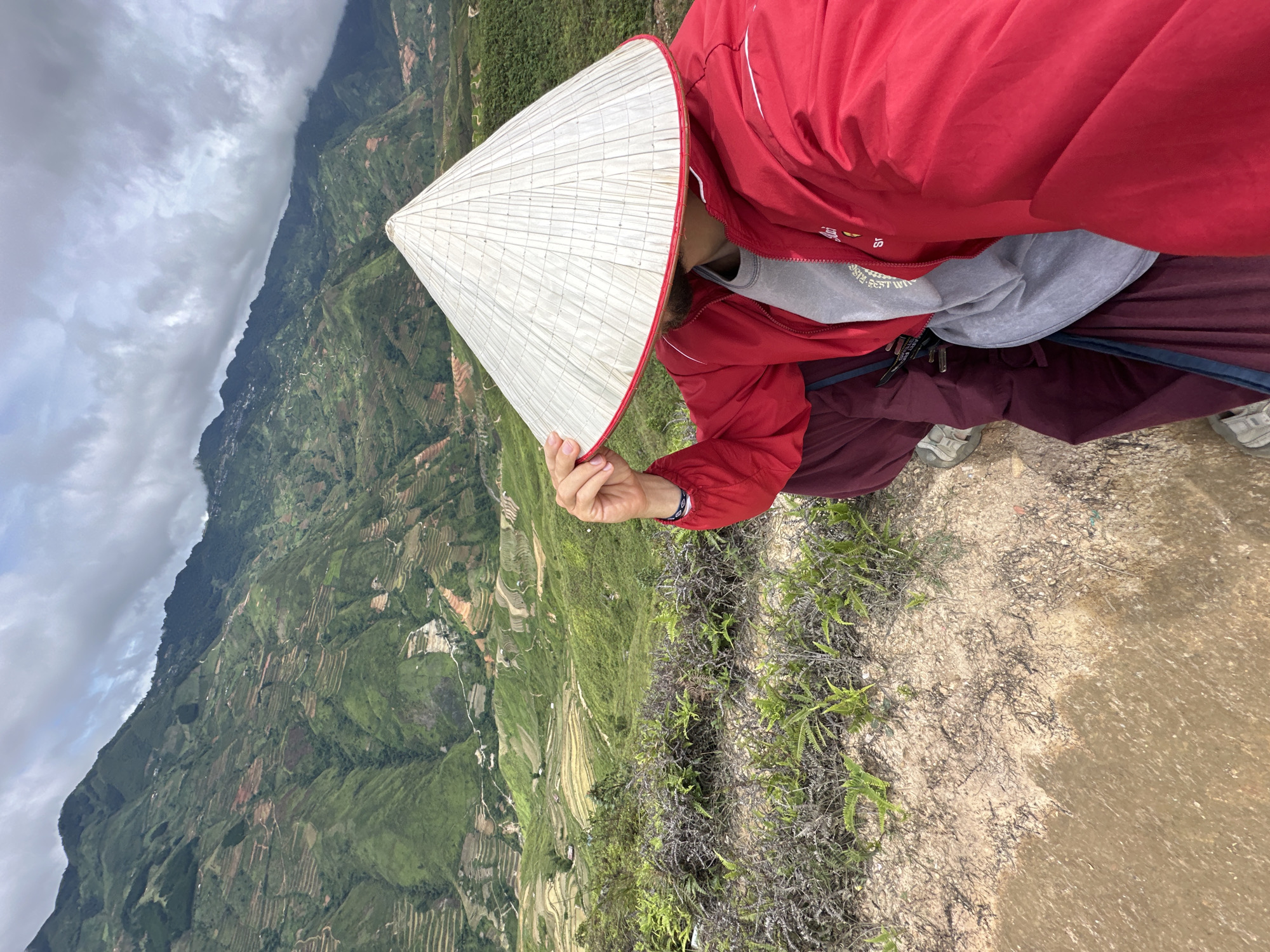 In Ta Xua, Vietnam, a person wearing a red jacket, maroon pants, and a conical hat sits on a dirt path overlooking a lush, green mountainous landscape. The mountains are terraced, with visible rice paddies and some scattered buildings. The sky is cloudy, with patches of sunlight breaking through. The person is holding the brim of the conical hat, obscuring their face. The scene captures a moment of repose amidst a scenic natural environment.