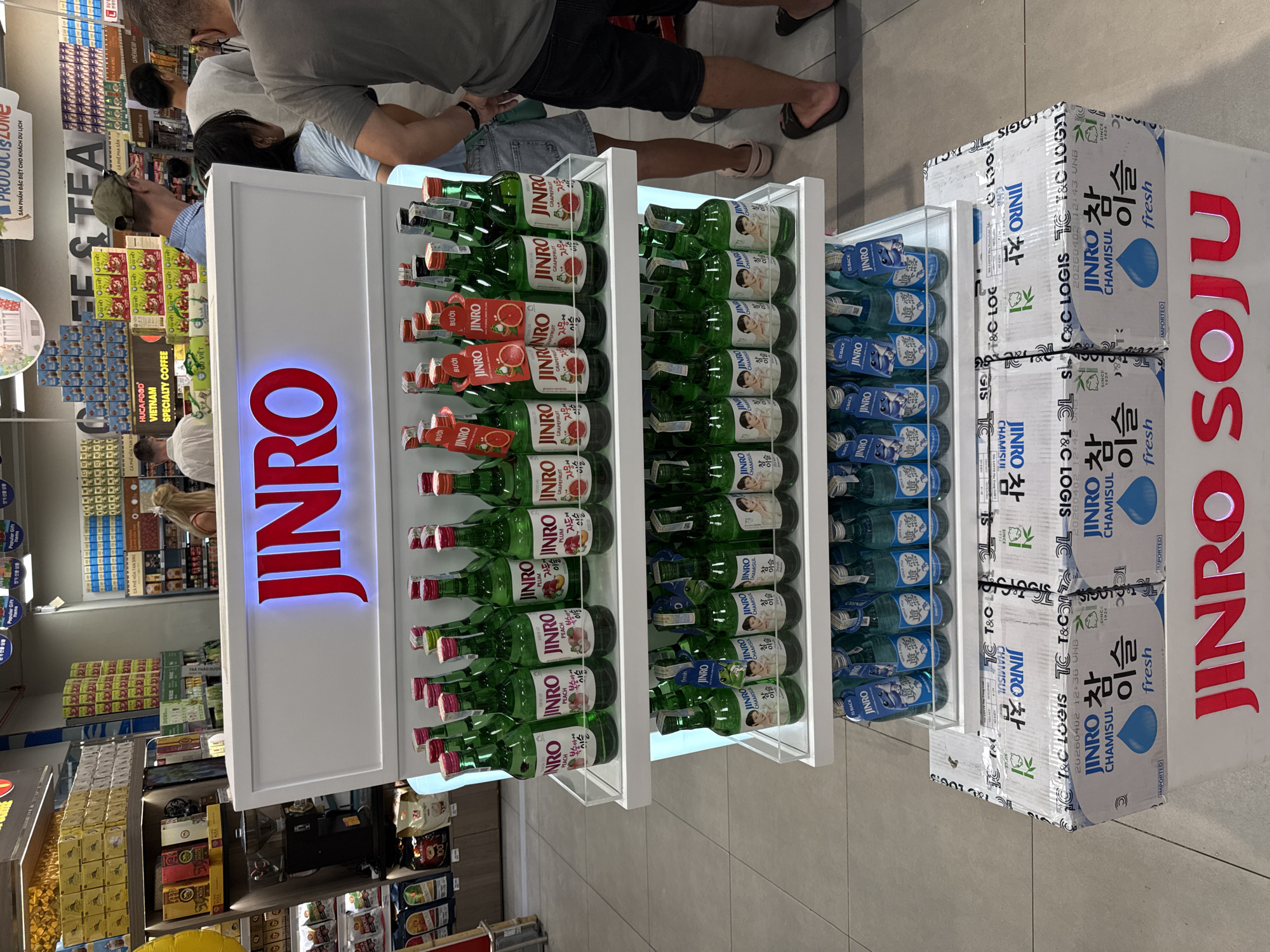 The image shows a retail display of Jinro Soju bottles in a store. The display features several shelves stocked with green bottles of soju, some with fruit-flavored labels (peach, plum, grapefruit) and others labeled "Chamisul Fresh." In the foreground, there are stacked boxes of Jinro Chamisul Soju, with the "Jinro Soju" branding prominently displayed on a white structure behind the bottles. In the background, shelves with various products, including coffee and tea, are visible. Several individuals are present in the store, some looking at the merchandise. The environment appears to be a well-lit supermarket or convenience store. The overall scene captures a product display in a commercial setting.