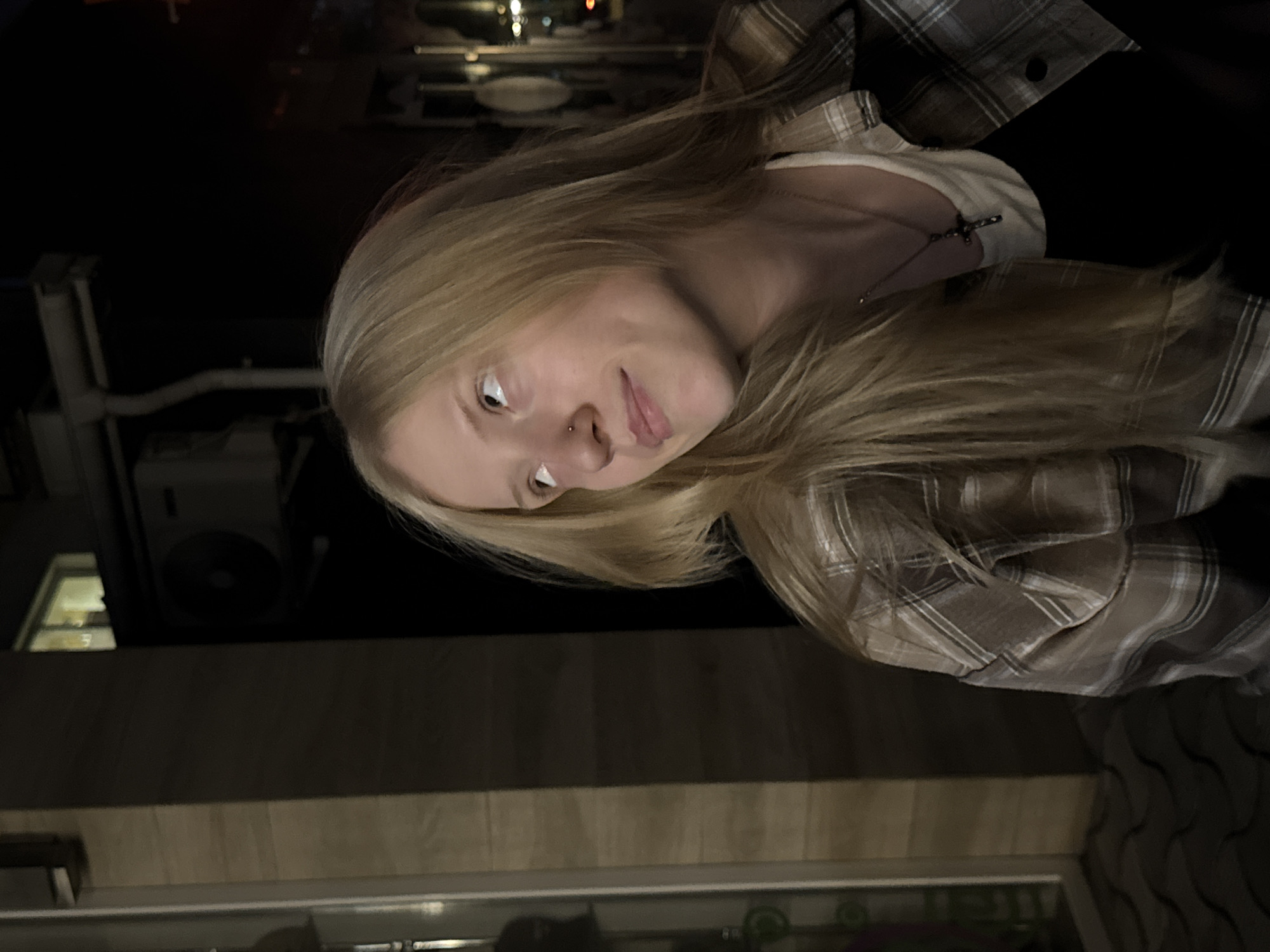 A young woman with long blonde hair and a nose ring looks up and to the right, her face illuminated by soft lighting. She is wearing a plaid shirt over a white tank top and a dark pendant necklace. The background is dark, suggesting it is nighttime, with hints of urban elements like a building with a window and an air conditioning unit. The scene appears to be outdoors, possibly on a street or in a courtyard in Tiraspol, Moldova.