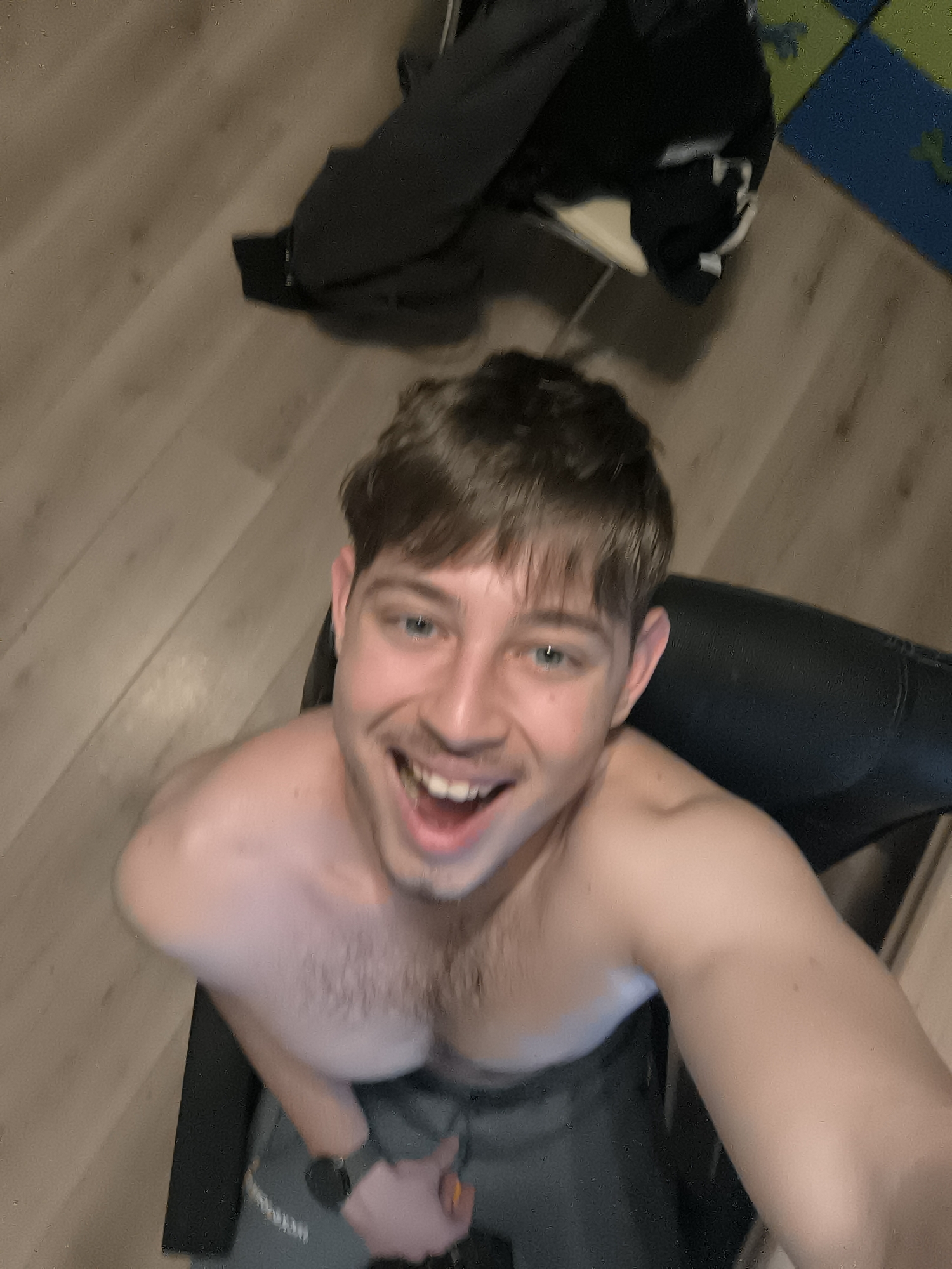A medium, high-angle shot shows a young, shirtless man sitting in a black chair. He is smiling with his mouth open and looking up at the camera. He is wearing gray pants and a black watch on his left wrist. A black jacket is on a chair behind him, with a blue and green playmat visible in the background. The floor is light brown wood. This image could not be pinpointed in the city of Sofia without more information.