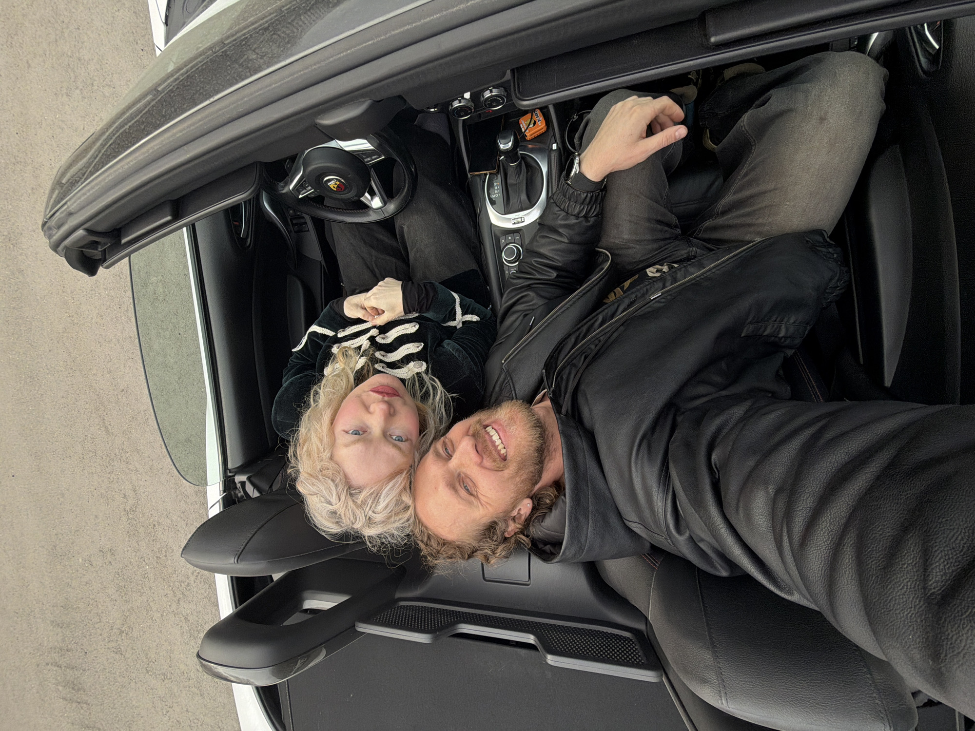 A low-angle, close-up photograph shows a couple in a car. The woman has blonde, curly hair and wears a black jacket with white stripes. The man has light brown, curly hair and a beard, and he wears a black leather jacket. Both are smiling and looking at the camera. The car's interior is visible, including the steering wheel, dashboard, and seats. The image provides no information about the city of Saint Petersburg, Russia.