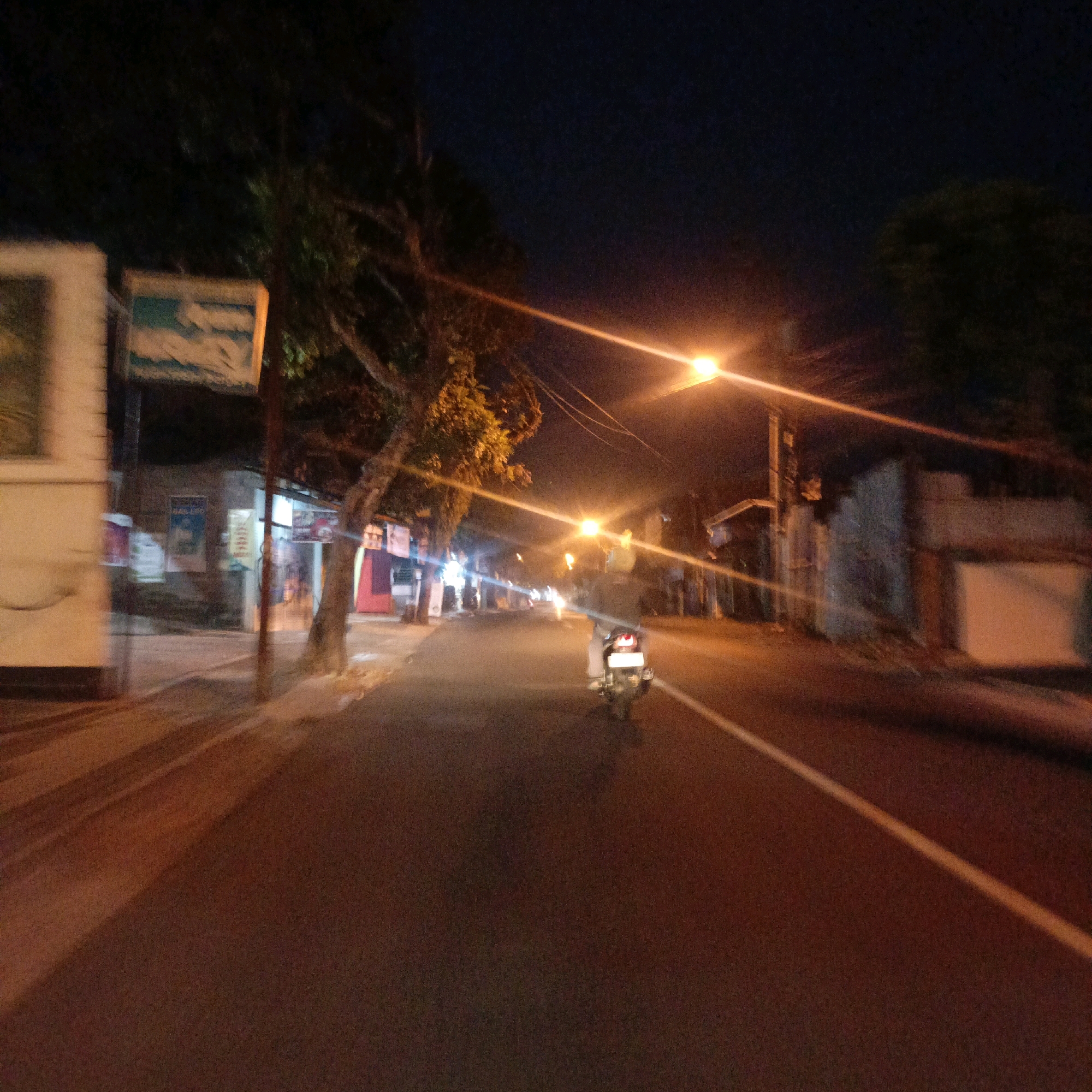 This is a nighttime, slightly motion-blurred photograph of a street in Salatiga, Indonesia. The image captures a road with buildings and trees lining one side. A single motorcycle is visible in the center of the road, moving away from the viewer. Streetlights illuminate the scene with warm, orange hues, casting long shadows across the asphalt. Some of the buildings appear to be shops, with signs and illuminated windows. The sky is dark, indicating it is nighttime.