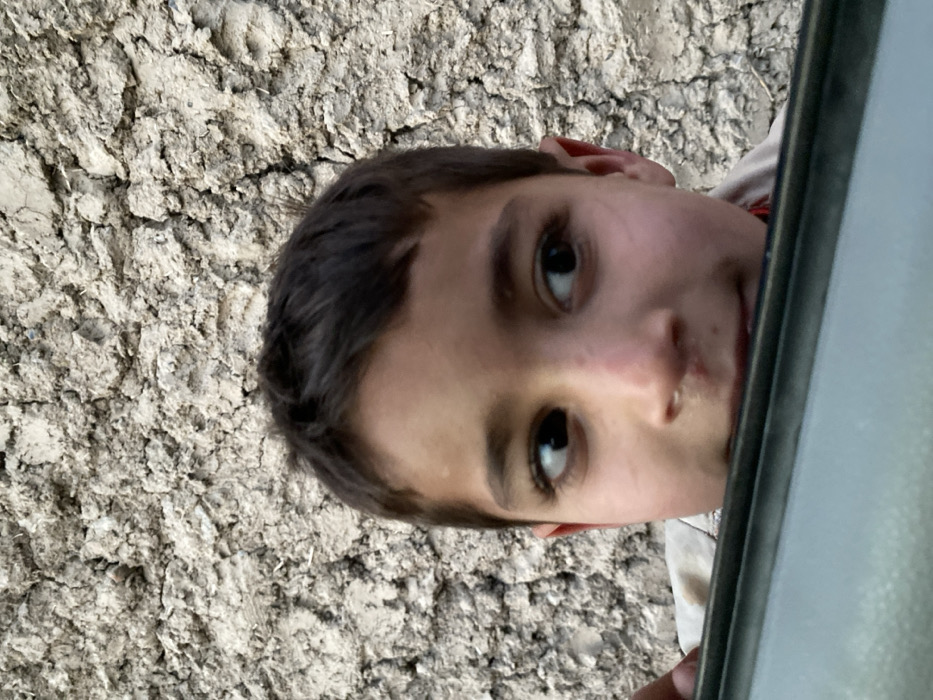 A close-up image captures a young male subject, estimated between 6-10 years of age, located in Kandahar, Afghanistan. The subject's head and upper chest are visible, positioned behind a dark, metallic horizontal object, consistent with the upper frame of a vehicle's door or window. He has dark brown hair and dark eyes, with a light complexion. His gaze is directed towards his left, and a subtle smirk is evident on his face. A portion of his left hand is visible gripping the metallic object on the far left of the frame. The background consists of a textured, light-colored wall, characteristic of mud or stucco construction, exhibiting numerous cracks and surface irregularities.