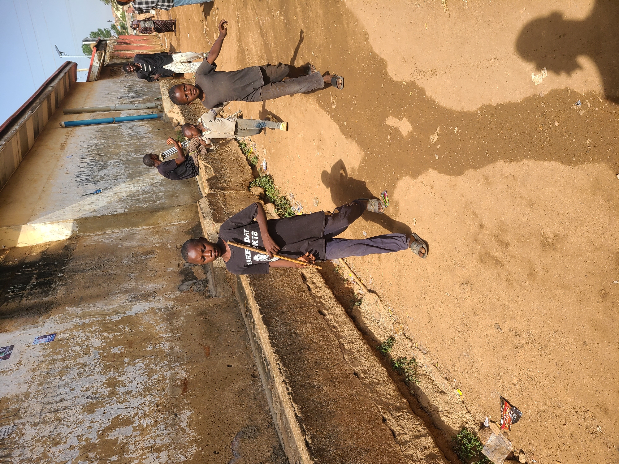 An outdoor scene in Jos, Nigeria, depicts several young males on an unpaved, sandy-brown path next to a concrete wall and drainage ditch under bright daylight.
In the foreground, a young boy in a black t-shirt with white text reading "MAKE DAY KIS" and dark pants stands, holding a long stick. He wears sandals and looks directly towards the camera with a neutral expression. To his left, another young boy in a black t-shirt and dark pants lies on his back on the ground with his arms outstretched. Near him, a third boy, dressed in a light-colored jacket over a t-shirt and light pants, appears to be sitting or crouching. Further back, near the weathered concrete wall, several other figures are visible; one in a black and white striped outfit and another in a dark shirt and light pants.
The ground is predominantly dry earth, with a few wet patches and scattered litter, including plastic debris and small pieces of paper. Patches of green vegetation grow along the edge of the concrete drainage ditch that runs parallel to the wall. The wall itself shows signs of wear and tear, with peeling paint and some indistinct graffiti or faded posters. In the background, part of a building with a red roof and a light pole are visible against a clear, bright sky. Strong shadows cast by the people and an unseen photographer suggest it is midday or early afternoon. The overall atmosphere appears casual, with the boys engaged in leisurely activity or resting.
