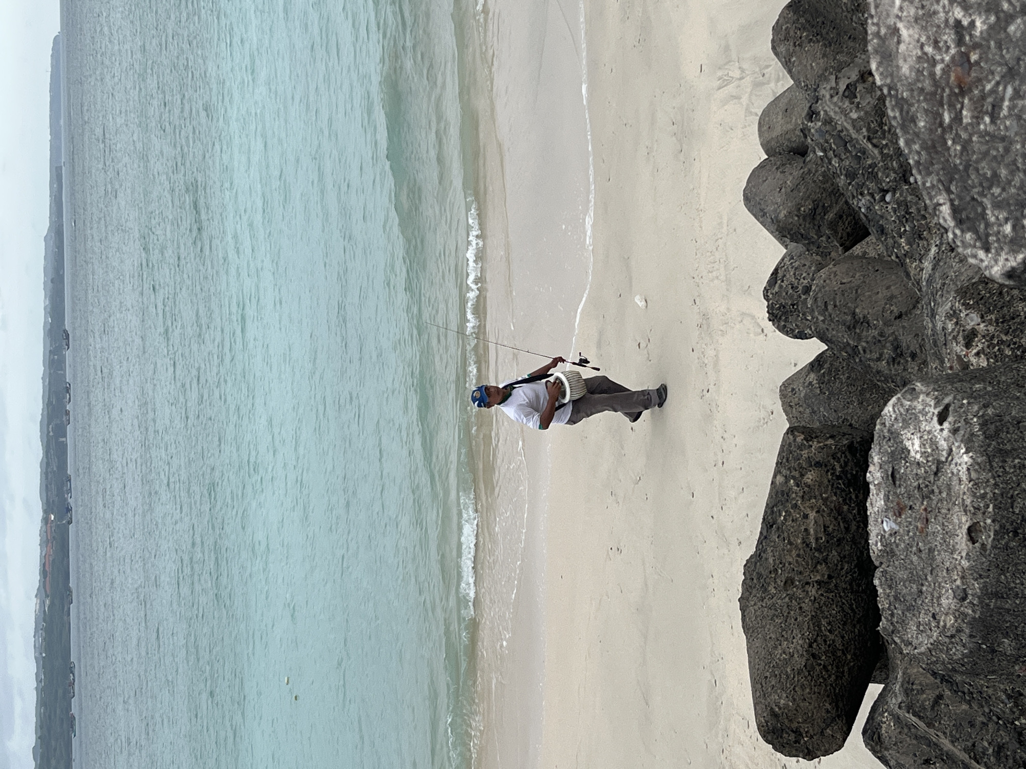 The image shows a man walking along a sandy beach in Kuta, Indonesia. He is wearing a white short-sleeved shirt, grey pants, and a blue cap. He is carrying a fishing rod in his right hand and a white bucket in his left hand. The sea is calm with gentle waves lapping at the shore. In the distance, several small boats are visible. In the foreground, large concrete blocks, possibly part of a breakwater or sea defense, are piled up. The sky is overcast.